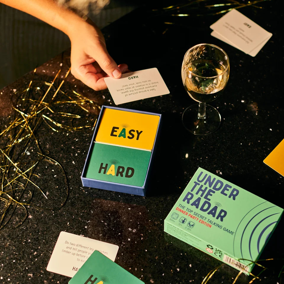 A hand places a small card into a blue box labeled EASY HARD, with two game boxes on a dark tabletop and a wine glass nearby. Visible product text includes EASY HARD and UNDER THE RADAR.