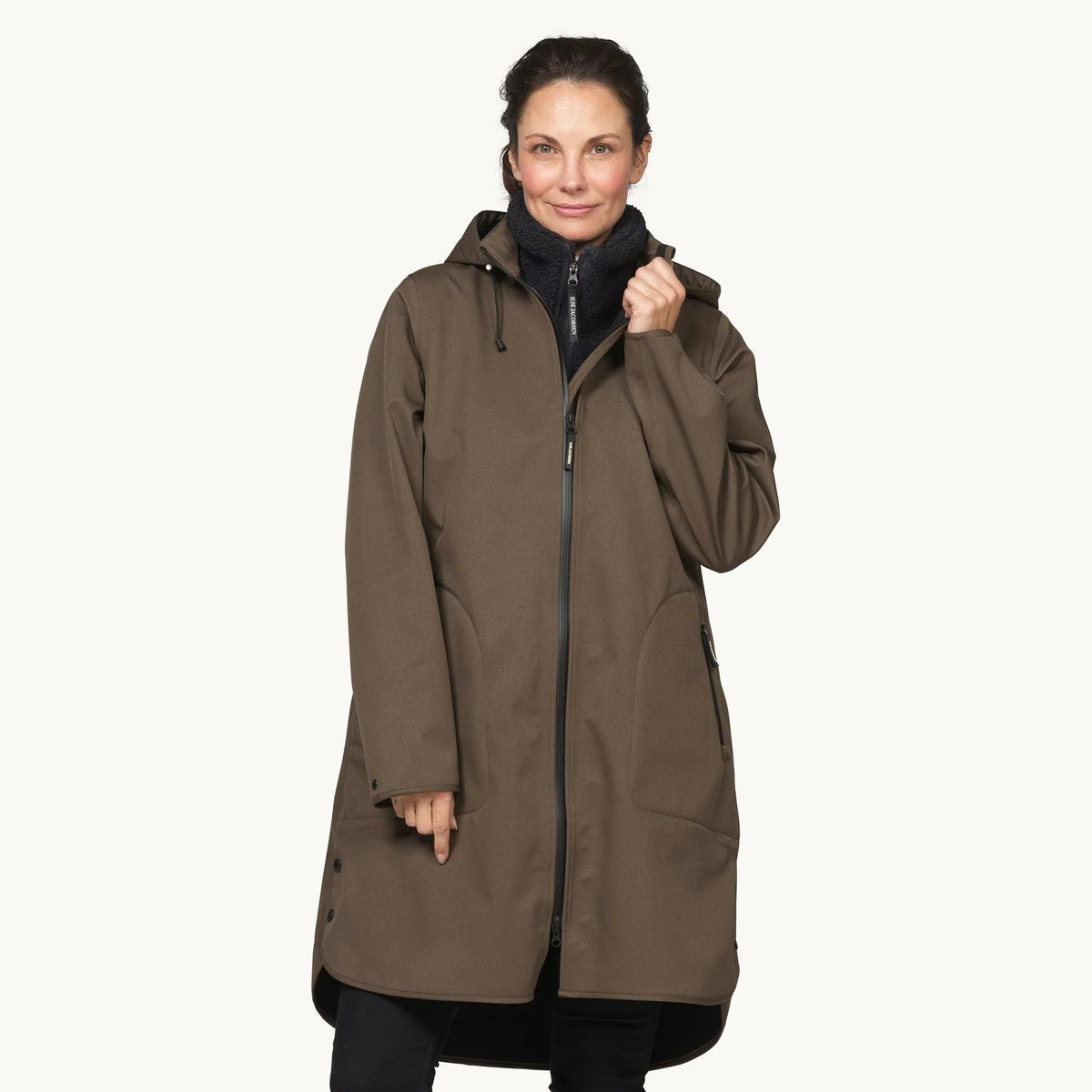 A woman modeling a mid-length brown hooded parka jacket with black zippers and black pants, standing against a plain off-white studio background.
