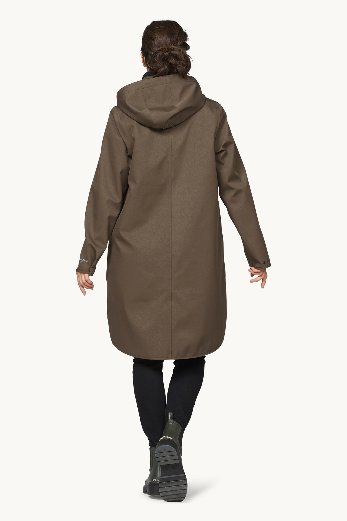 A woman viewed from behind wearing a mid-length hooded coat in a muted taupe-brown color, paired with black leggings and dark boots, against a plain off-white studio background.