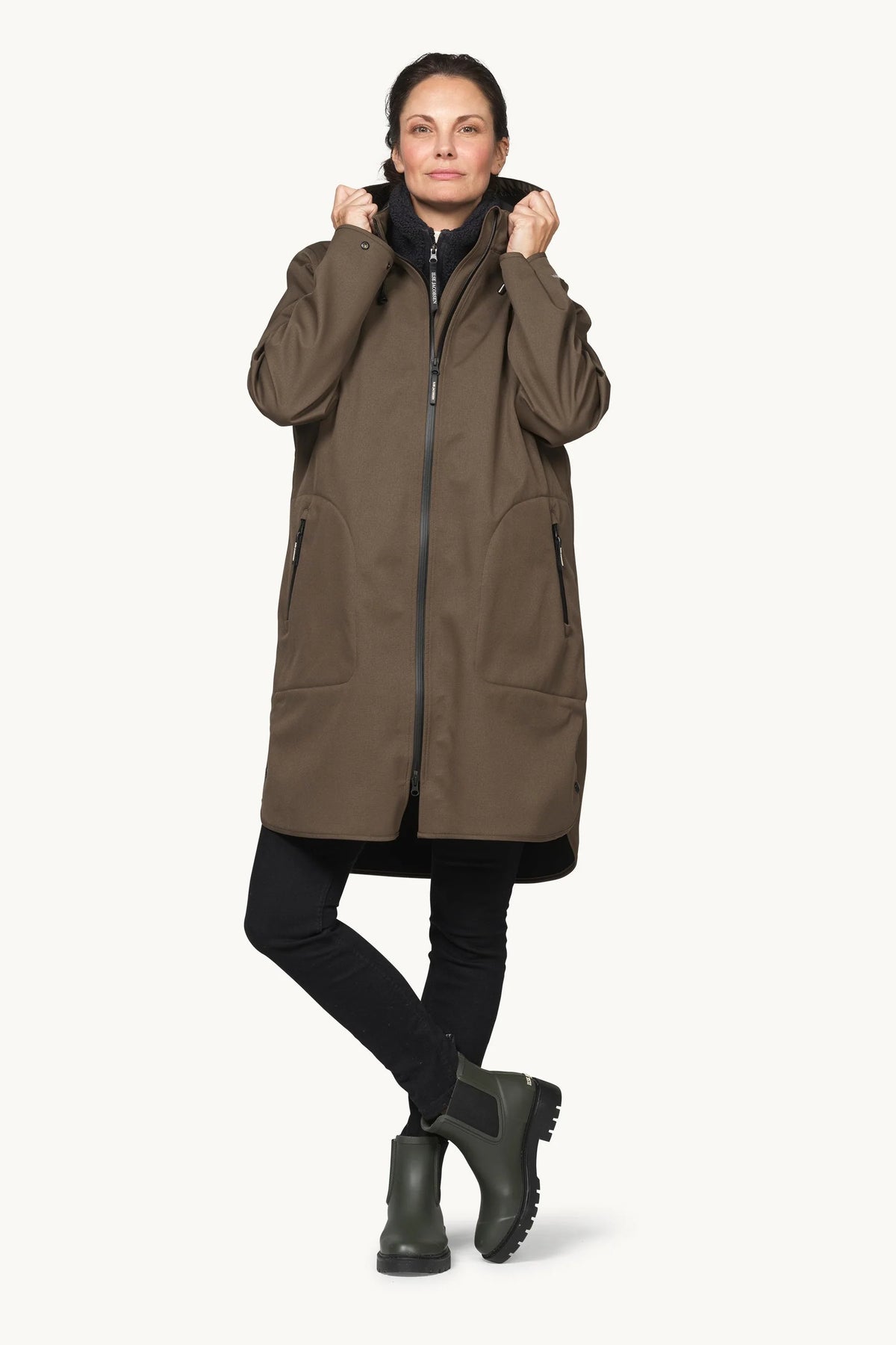 A woman modeling a mid-length olive brown hooded jacket with a front zipper, black pants, and dark green boots against a plain off-white background.