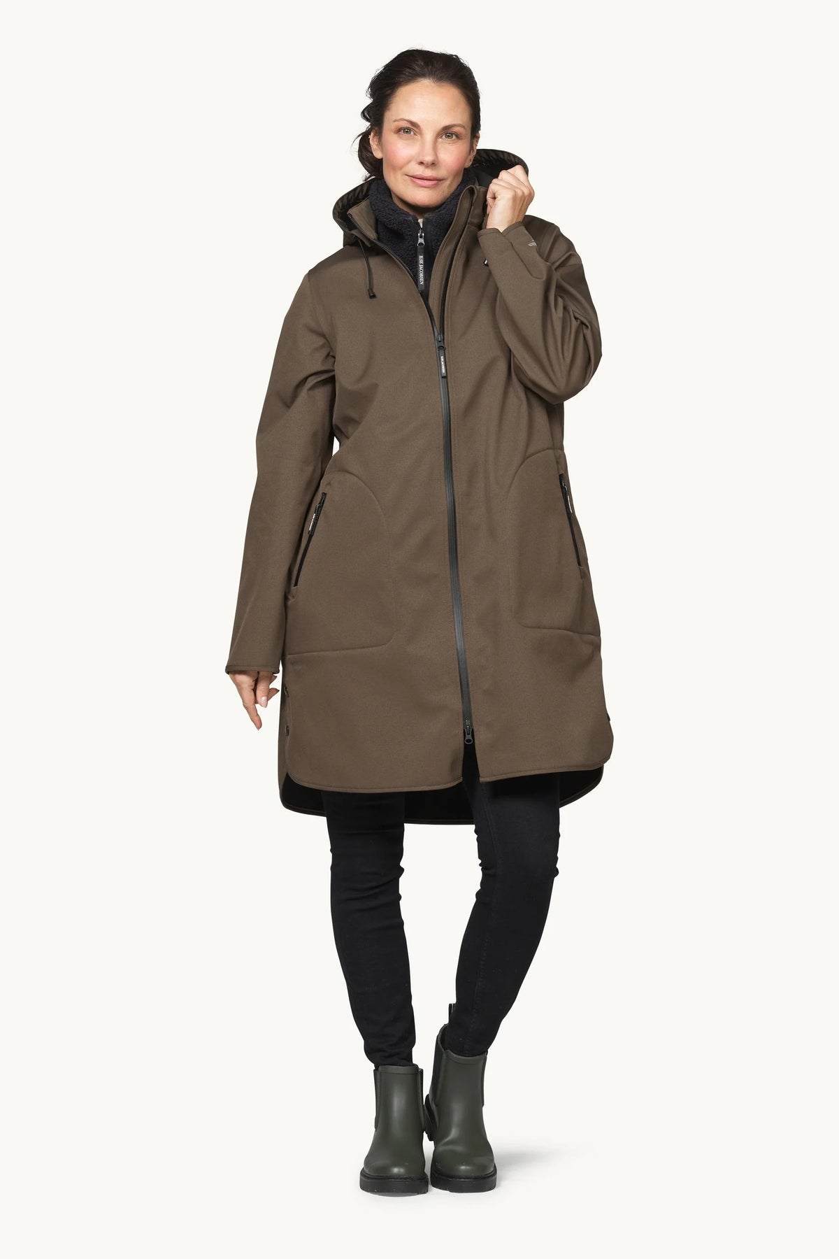 A woman modeling a mid-length brown hooded jacket with a front zipper, black zipper details, and side pockets, paired with black pants and dark rain boots, against a plain off-white background.