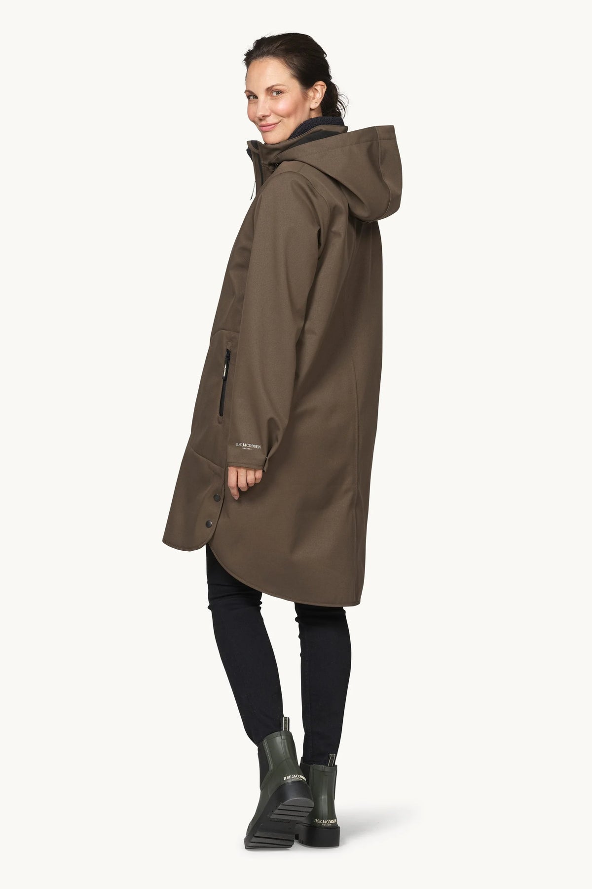 A woman modeling a mid-length hooded parka jacket in a dark brownish-olive color, paired with black leggings and dark green rain boots, against a clean off-white background.