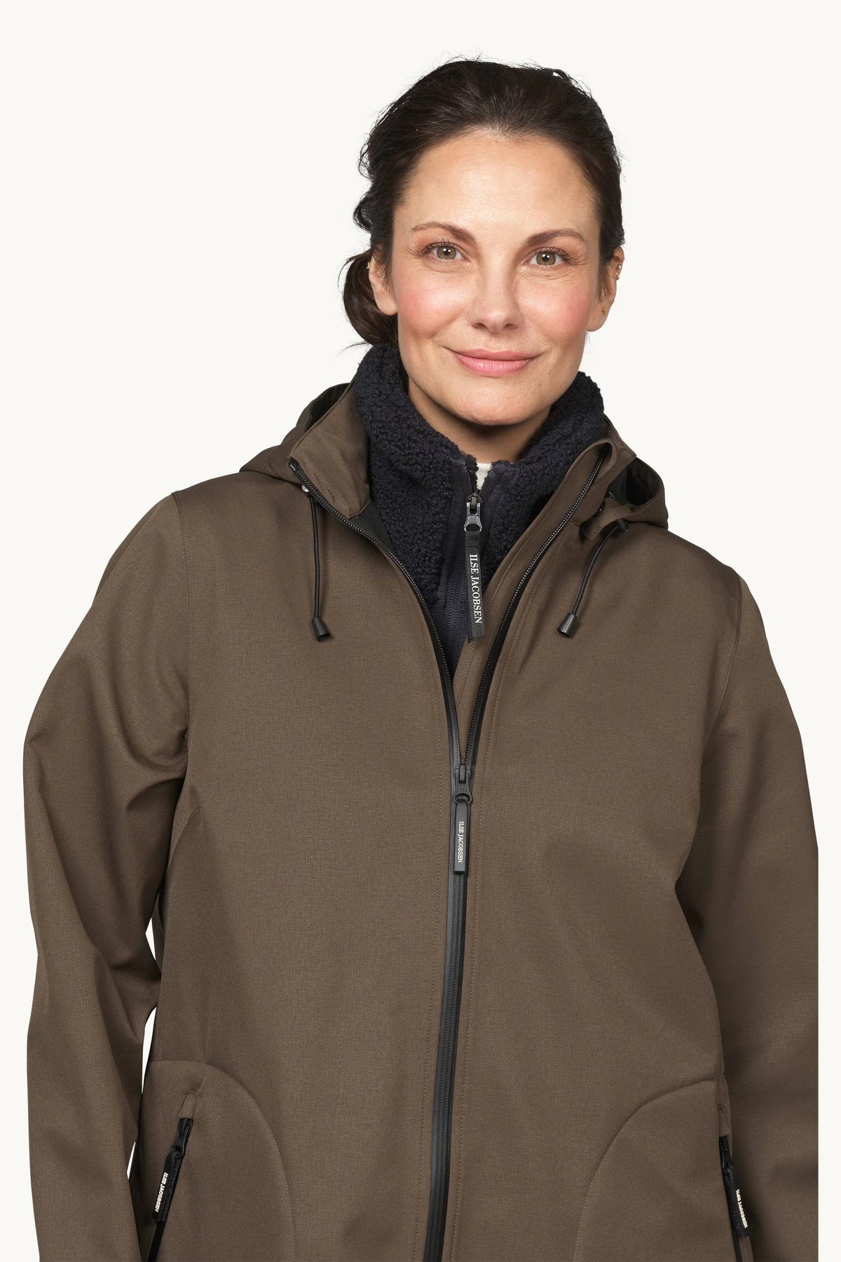 A woman wearing a brown hooded zip-up jacket over a dark fleece neck warmer, photographed against a plain light background.