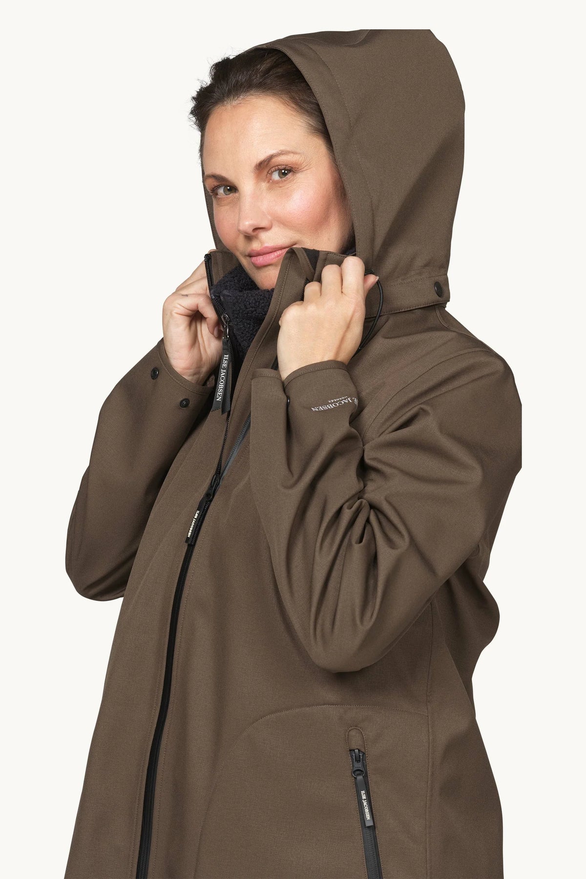 A woman wearing a hooded waterproof jacket in a warm brown color, shown from the chest up against a light off-white background.