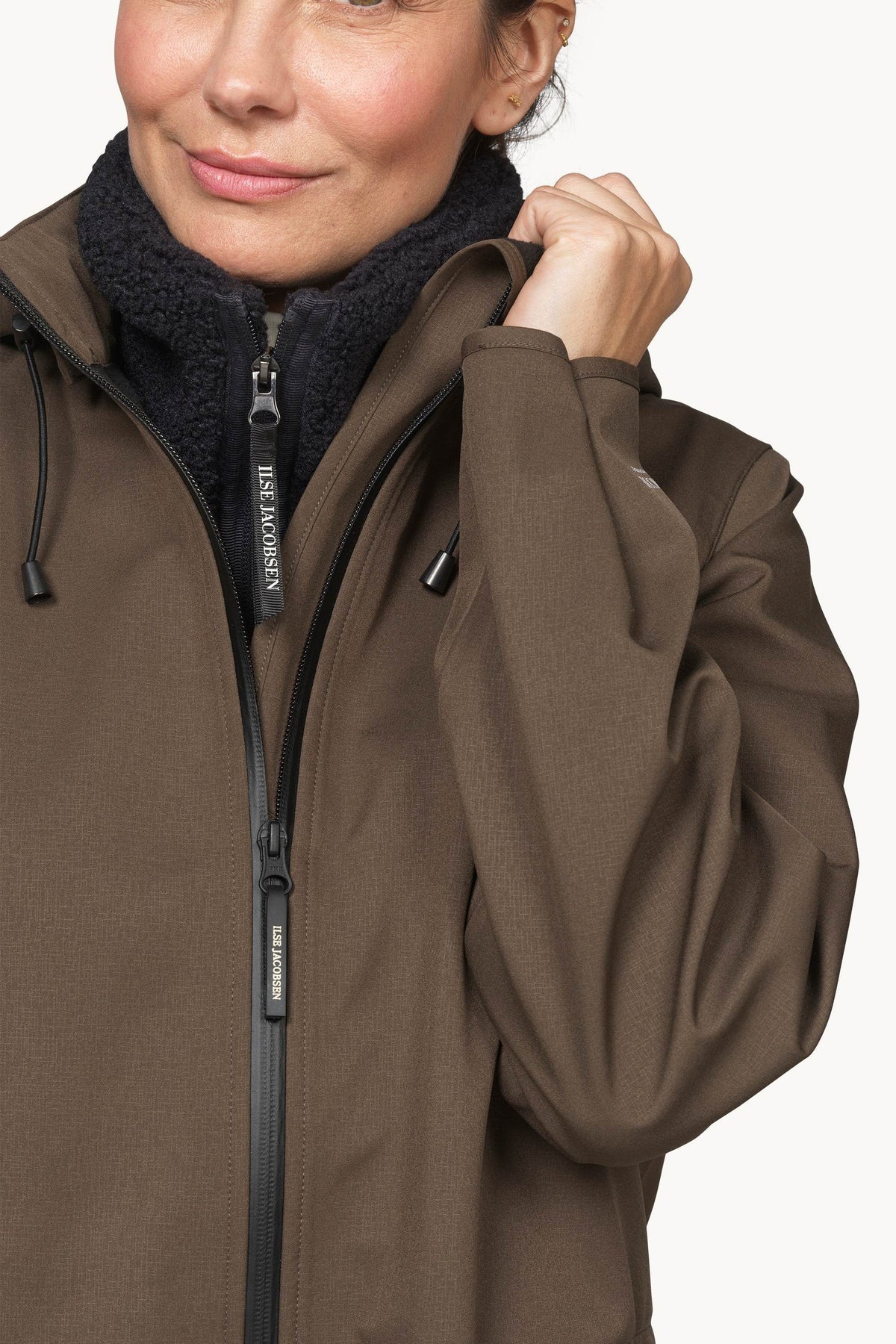 A close-up of a woman wearing a brown waterproof jacket with a black fleece collar and a black zipper, holding up the collar area. The brand name 'HELM HANSEN' is visible on the zipper pull.