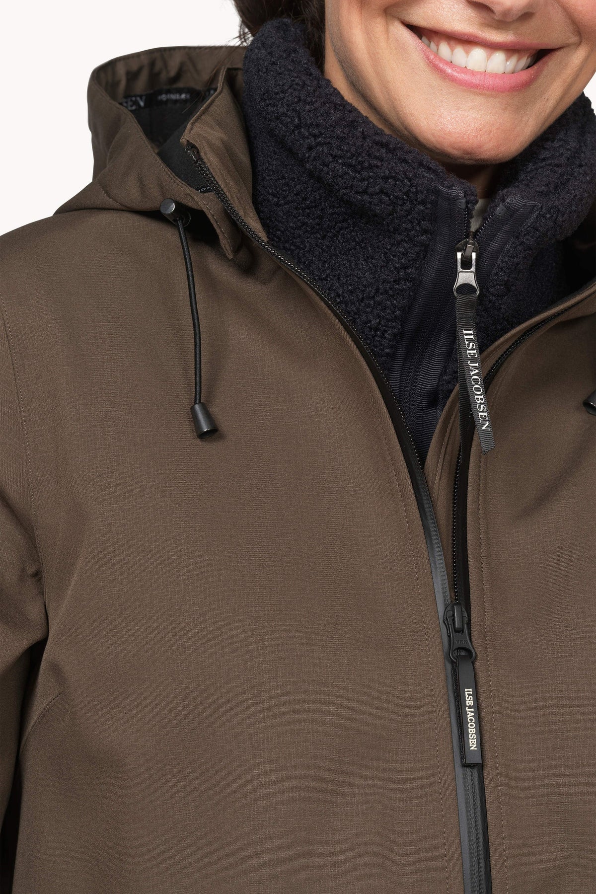 Close-up of a person wearing a brown hooded jacket with a dark fleece collar and black zipper, against a light studio background.