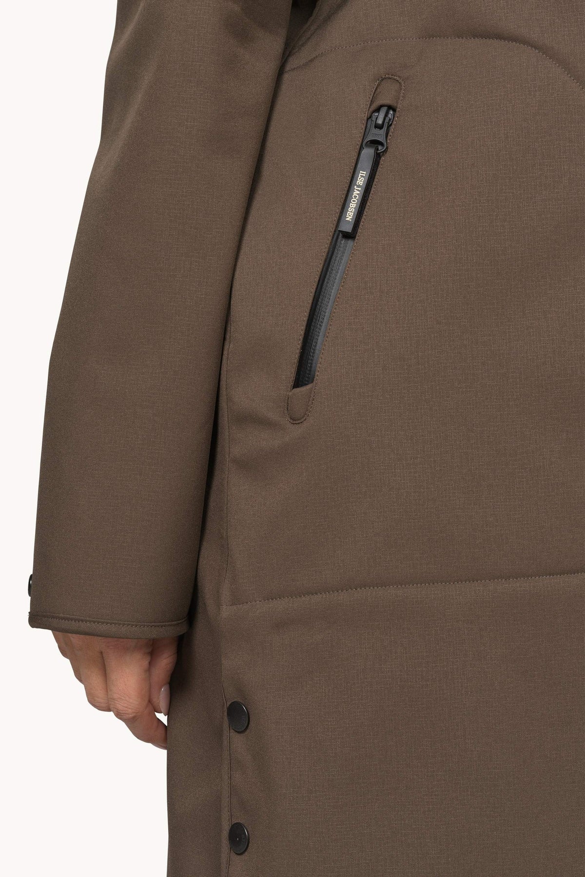 Close-up of a brown outerwear jacket with a vertical zip pocket and black buttons, shown on a model against a plain light background.