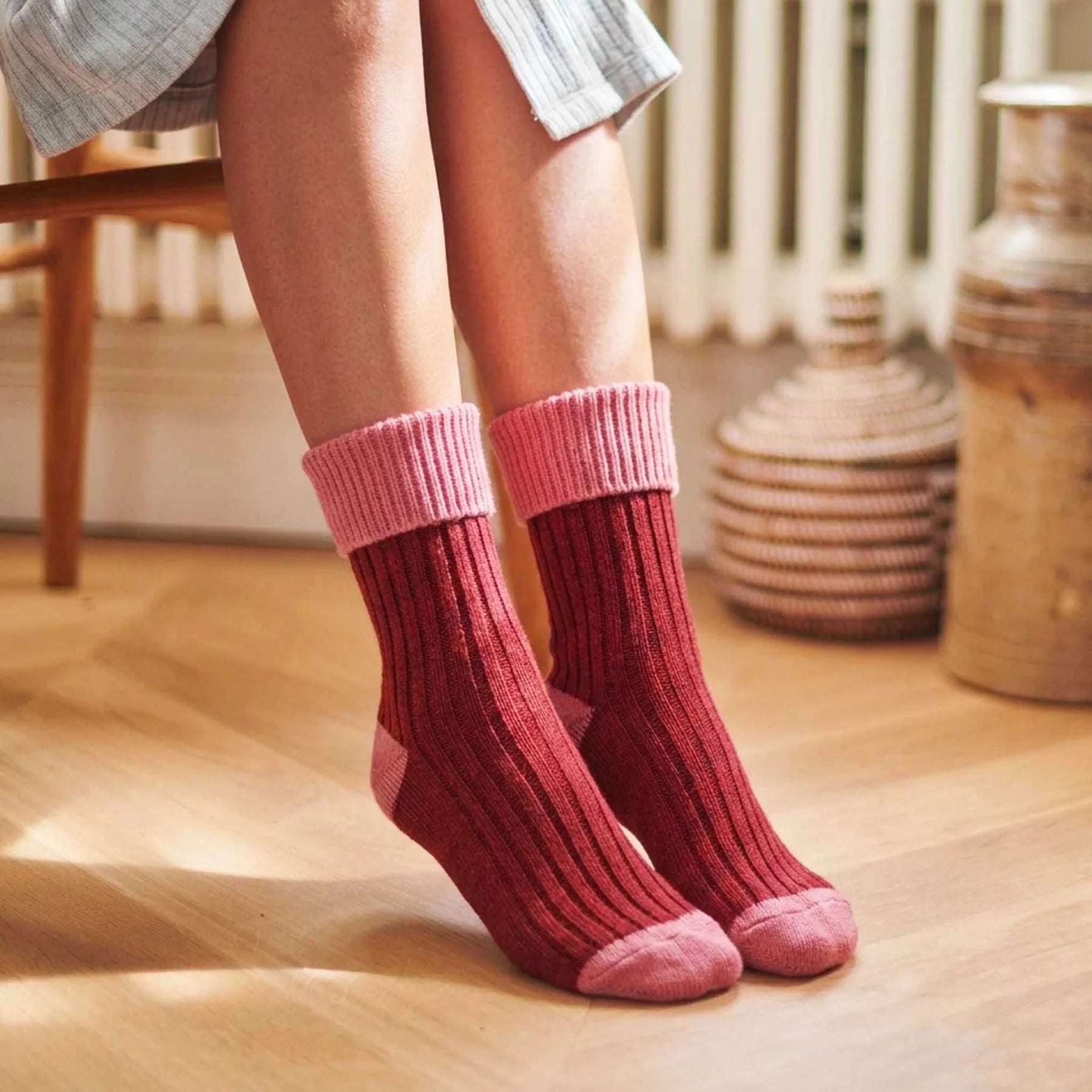 A person wearing a pair of ribbed crew socks in dusty rose red with lighter pink cuffs and toes, shown in a warm indoor setting on a wooden floor.