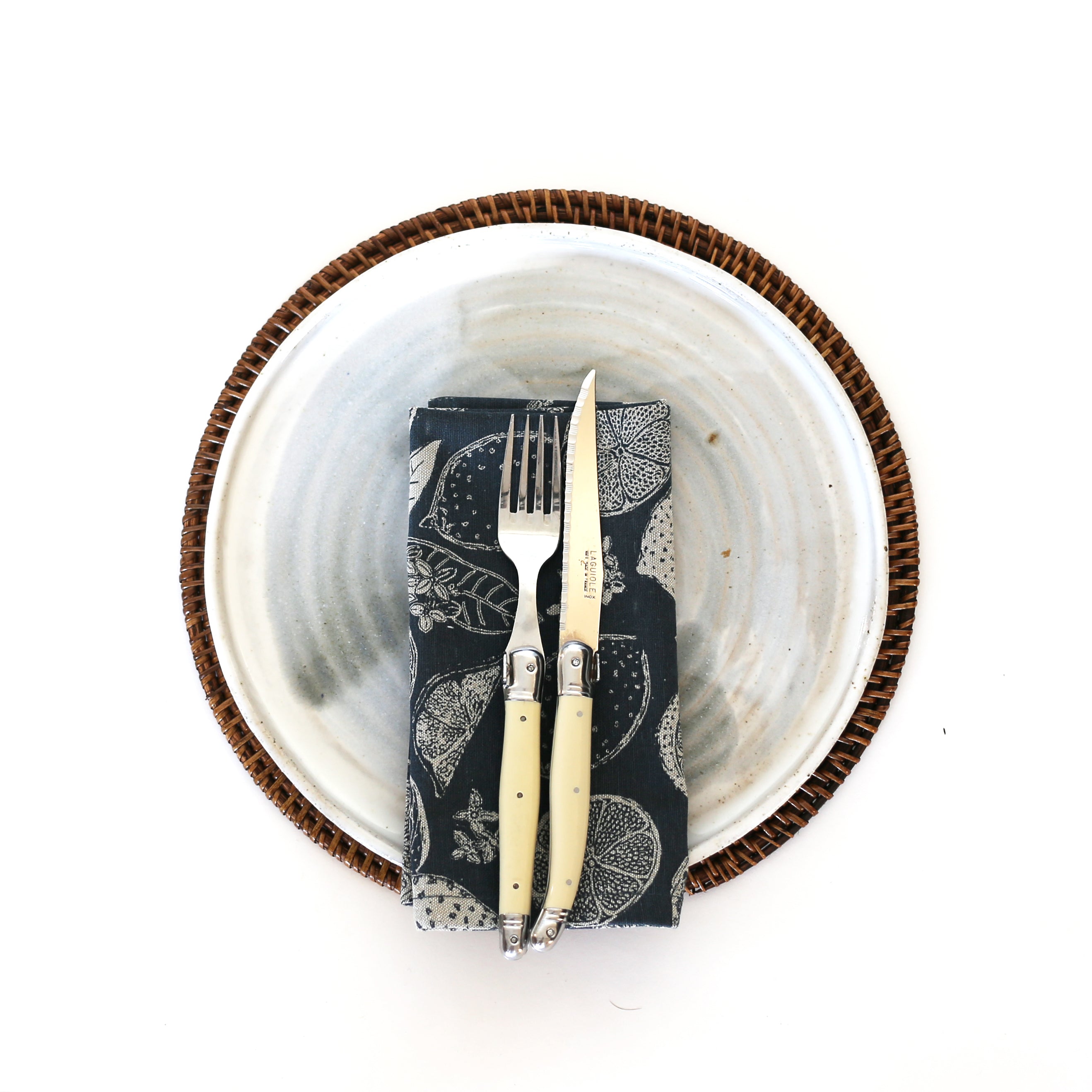 A round, speckled white plate with a brown wicker charger, featuring a knife and fork with cream handles on a dark napkin