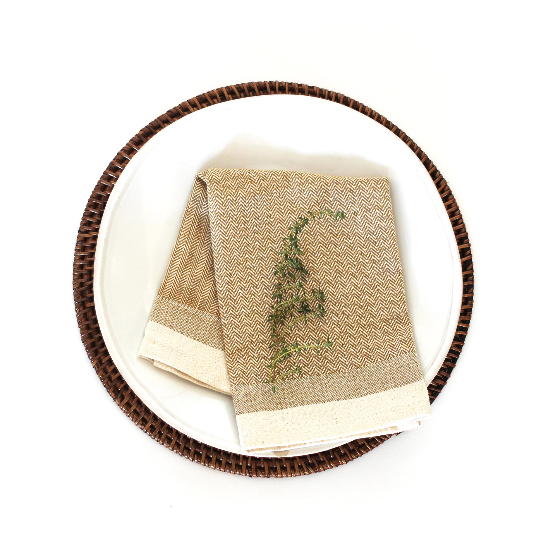 Mustard yellow herringbone napkin with off-white border, folded on a white plate with a dark brown woven charger.