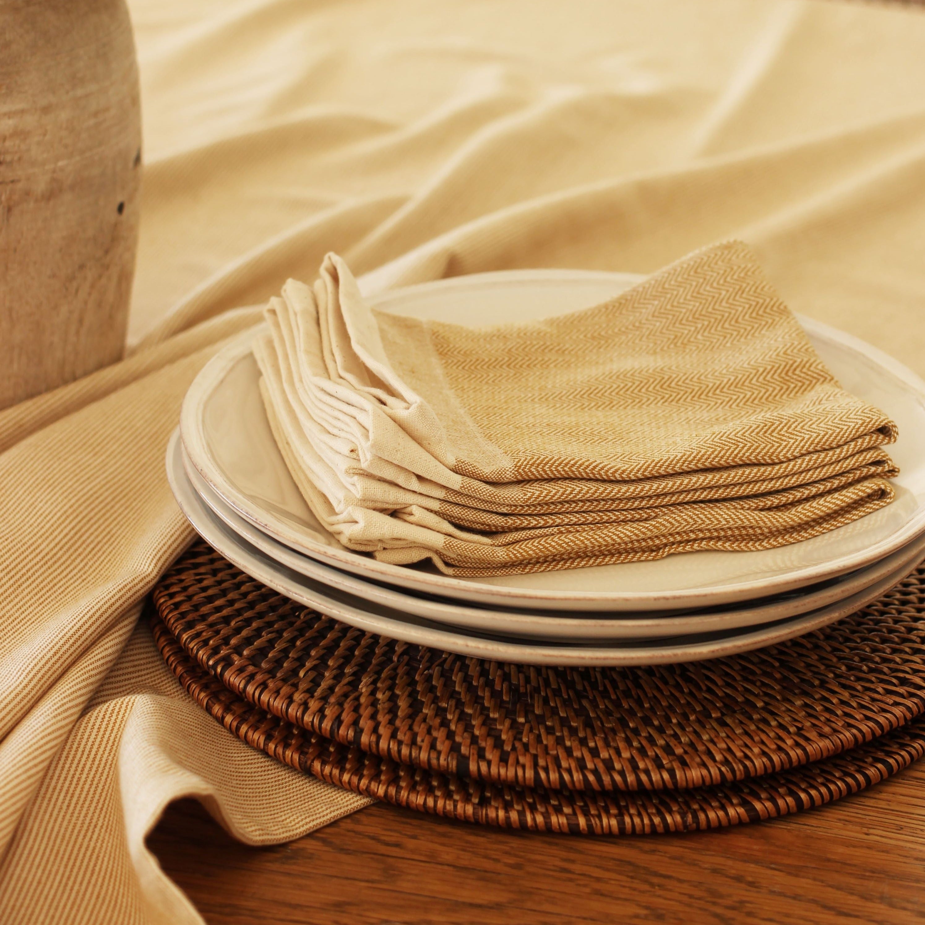 Stack of beige and gold herringbone napkins on top of stacked off-white plates, all resting on woven placemats.