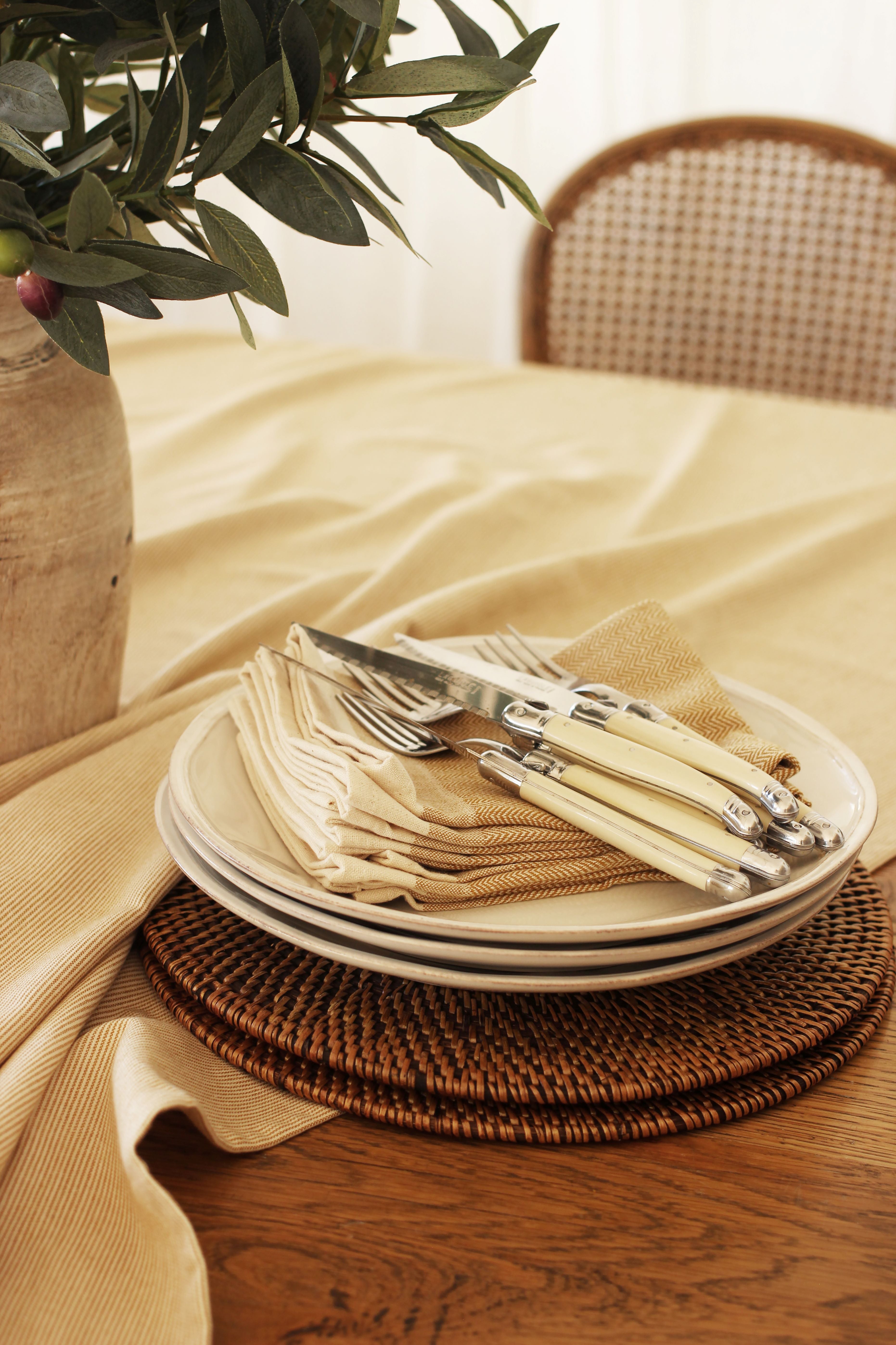 Stack of white plates with ivory-handled cutlery and beige napkins, resting on woven placemats.