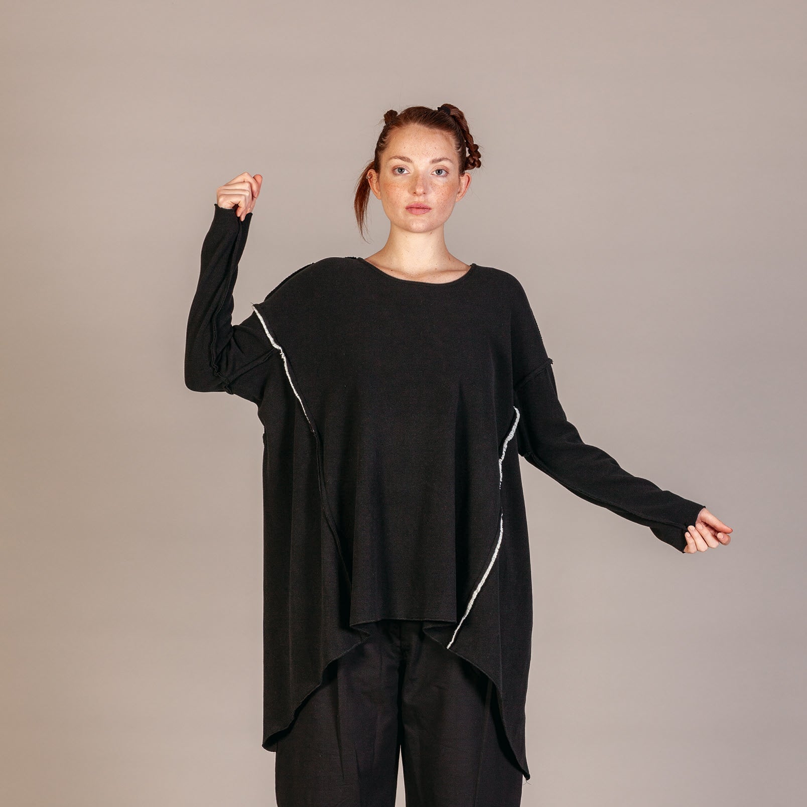A model wearing a black oversized long-sleeve top with white piping details, black cropped pants, and black-and-white striped socks, posed against a neutral gray studio background.
