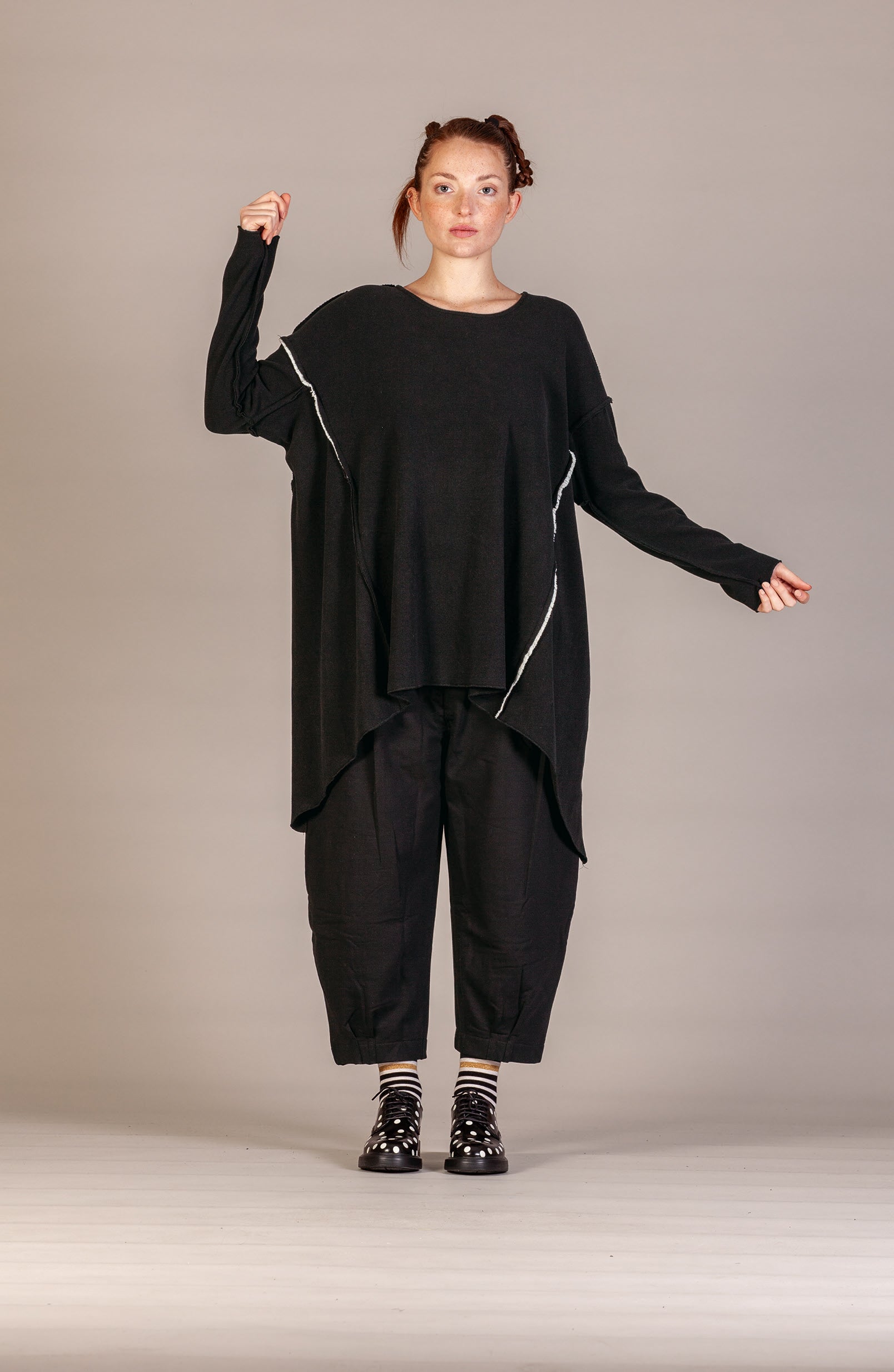A model wearing a black oversized long-sleeve top with white piping details, black cropped pants, and black-and-white striped socks, posed against a neutral gray studio background.