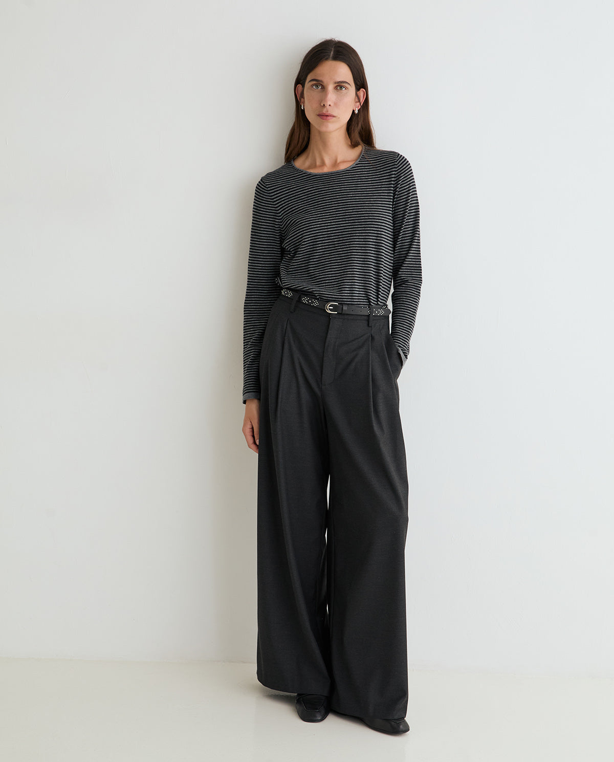 A woman stands against a plain white wall wearing a dark gray and black horizontally striped long-sleeve top with black wide-leg trousers and a thin black belt.