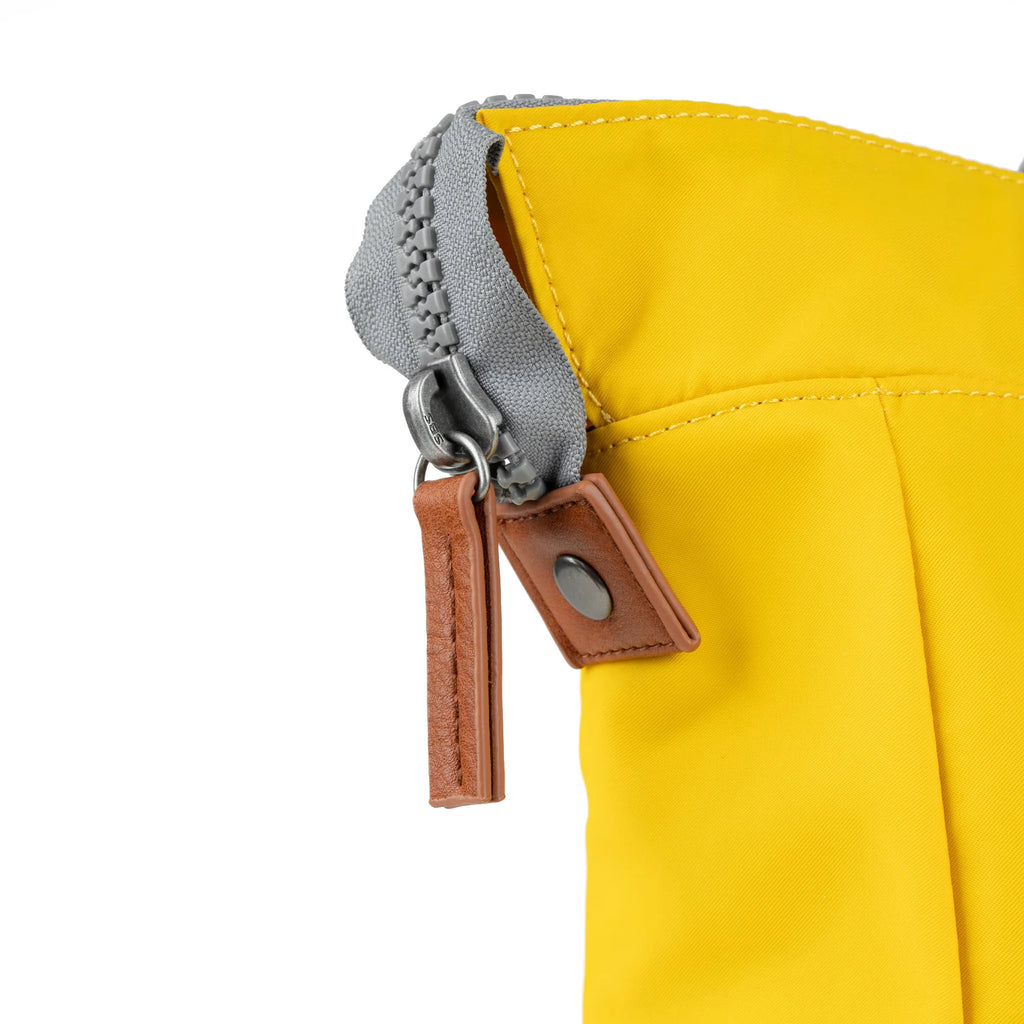 Close-up of a mustard yellow bag's zipper detail. The zipper is grey with a brown leather pull and tab.