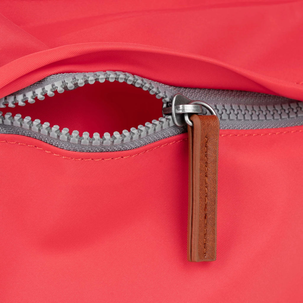 Close-up of a ROKA bag zipper, featuring a light gray zipper and a leather pull tab on a coral background.