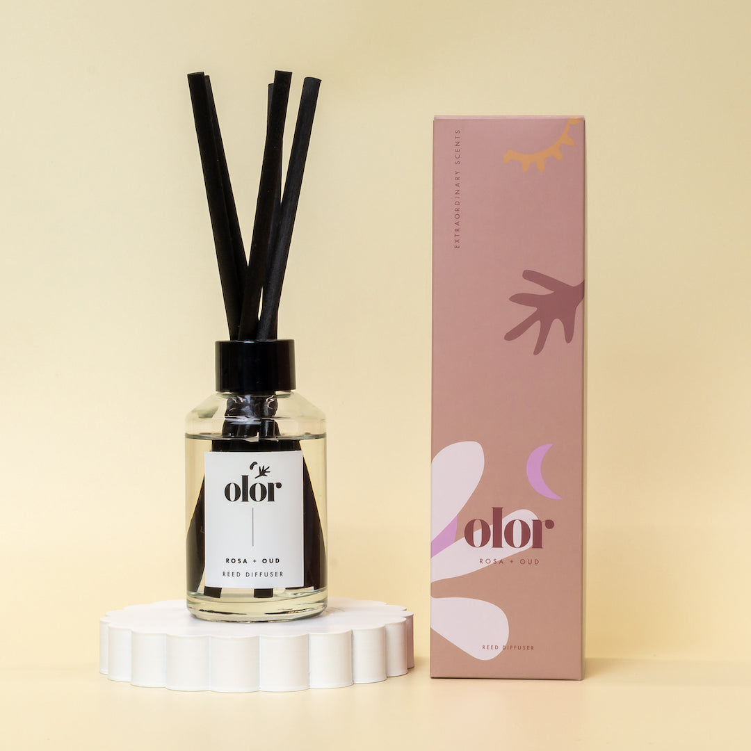 Olor Rosa + Oud reed diffuser in clear glass bottle with black reeds, shown with its packaging.