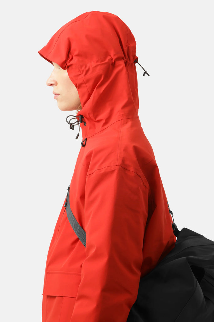 Profile view of a person wearing a bright red waterproof jacket with a drawstring hood and a black bag slung over their shoulder.