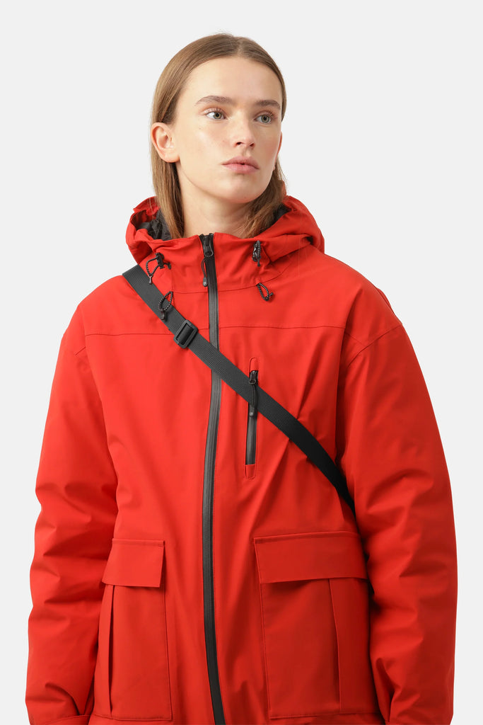Woman wearing a bright red hooded waterproof jacket with black zipper and pockets; a black strap crosses her body.