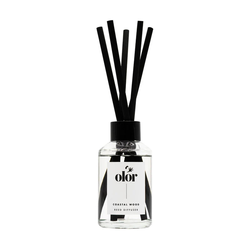 Clear glass reed diffuser bottle with black reeds and a black label reading "olor" and "Coastal Wood Reed Diffuser".