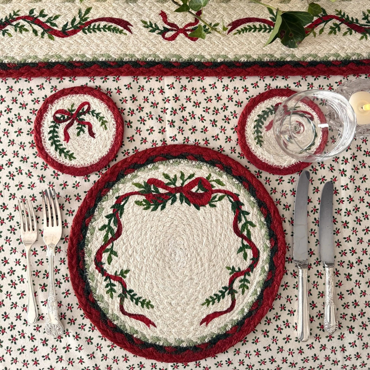 A festive holiday table setting featuring a braided cream placemat with red and green embroidered holly and bow motifs, matching coasters, a dinner plate, and flatware on a patterned tablecloth under green ivy and warm string lights.