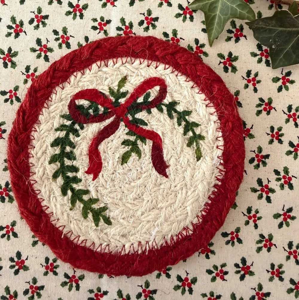 A round printed Christmas wreath patch with a red border, green holly leaves, and a red bow on a cream background, placed on a red and green holly patterned fabric with ivy leaves.
