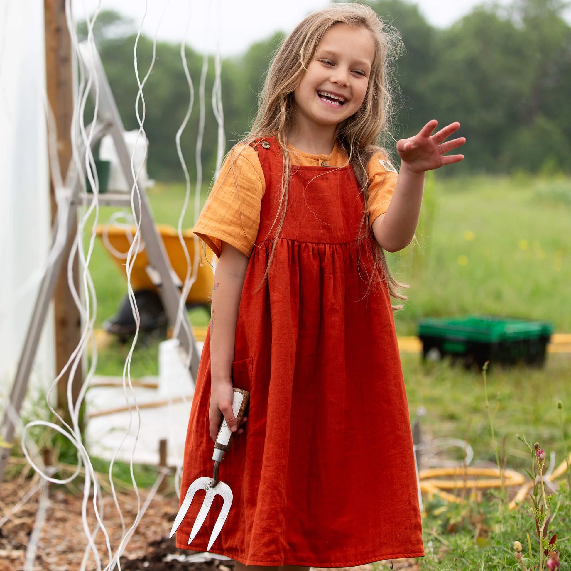 Young girl in a red dress holding a garden fork, standing in a garden.