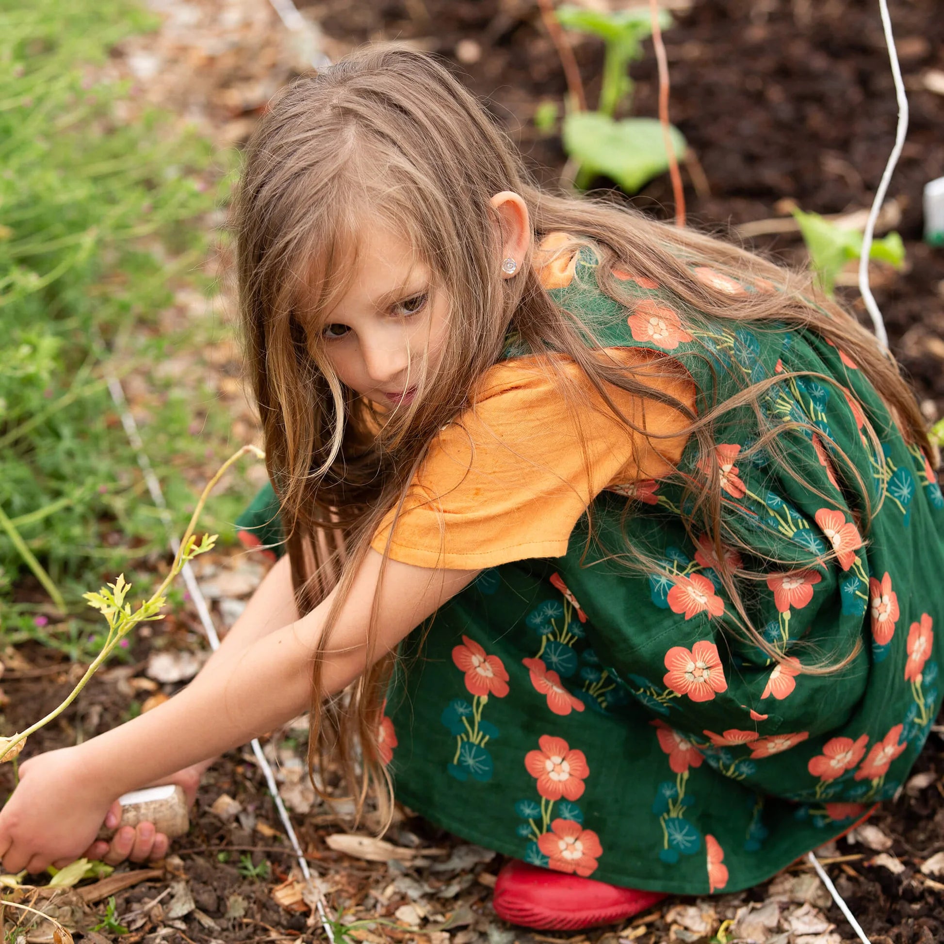 Young girl in a garden wearing a green floral dress and orange shirt.