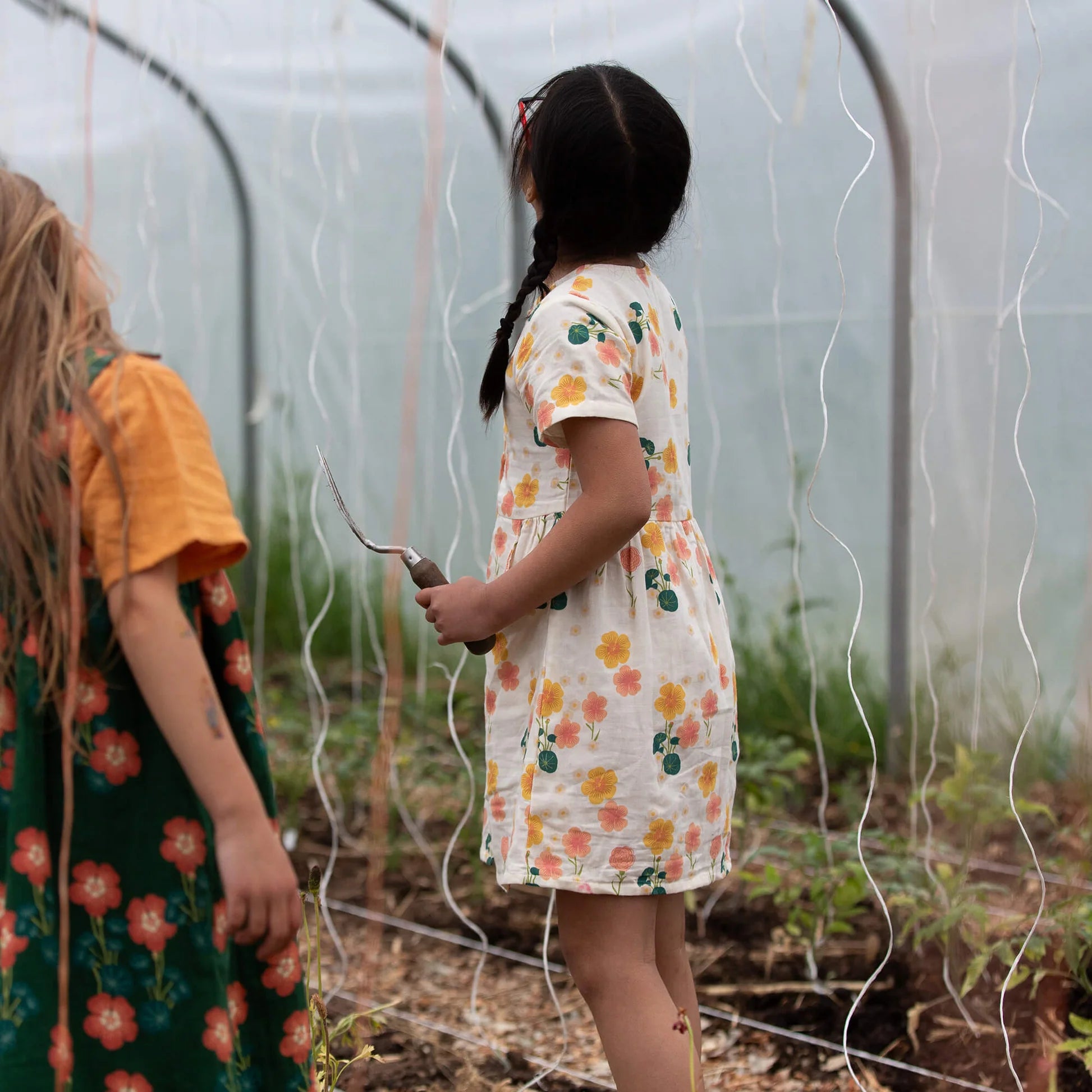 Two children in floral dresses standing inside a greenhouse.