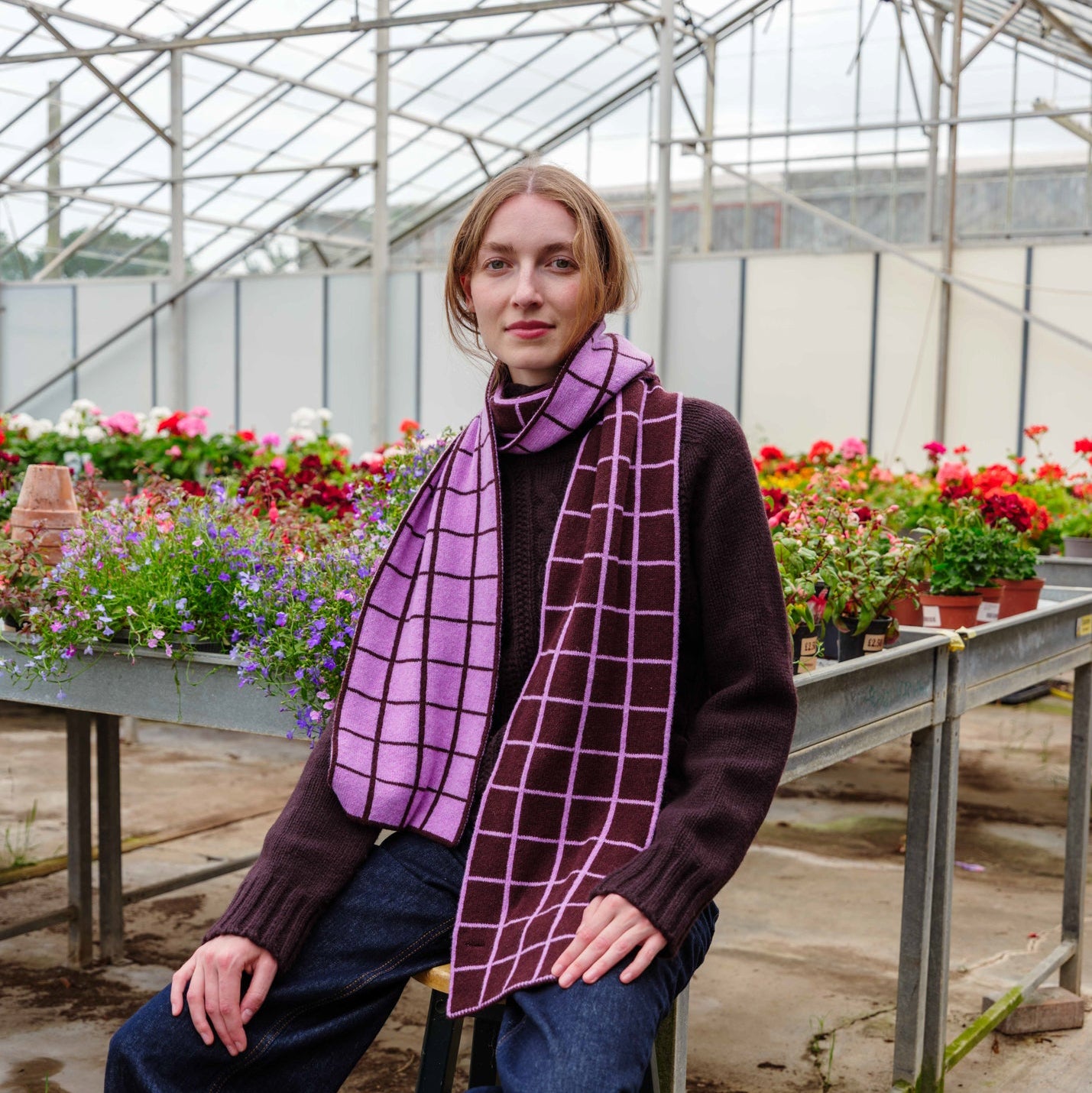 A woman sits on a stool inside a greenhouse, wearing a dark brown sweater, blue jeans, and a purple scarf with a pink grid pattern.