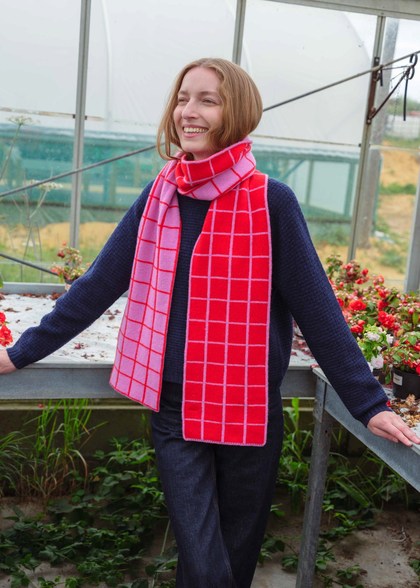 A woman wearing a red and pink plaid scarf, a navy blue sweater, and dark jeans stands in a greenhouse, smiling and looking to the side.