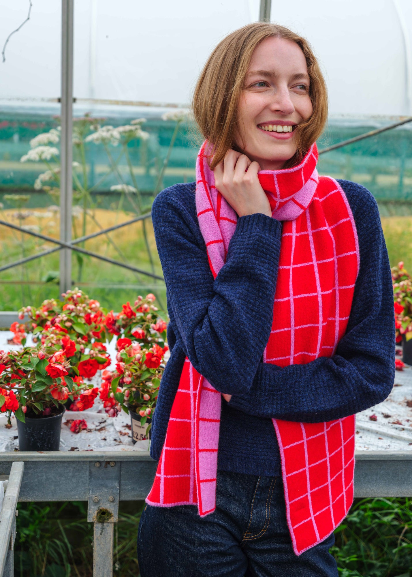 A woman wearing a navy blue knit sweater and a bright red and pink plaid scarf, standing in a greenhouse with potted red flowers in the background.