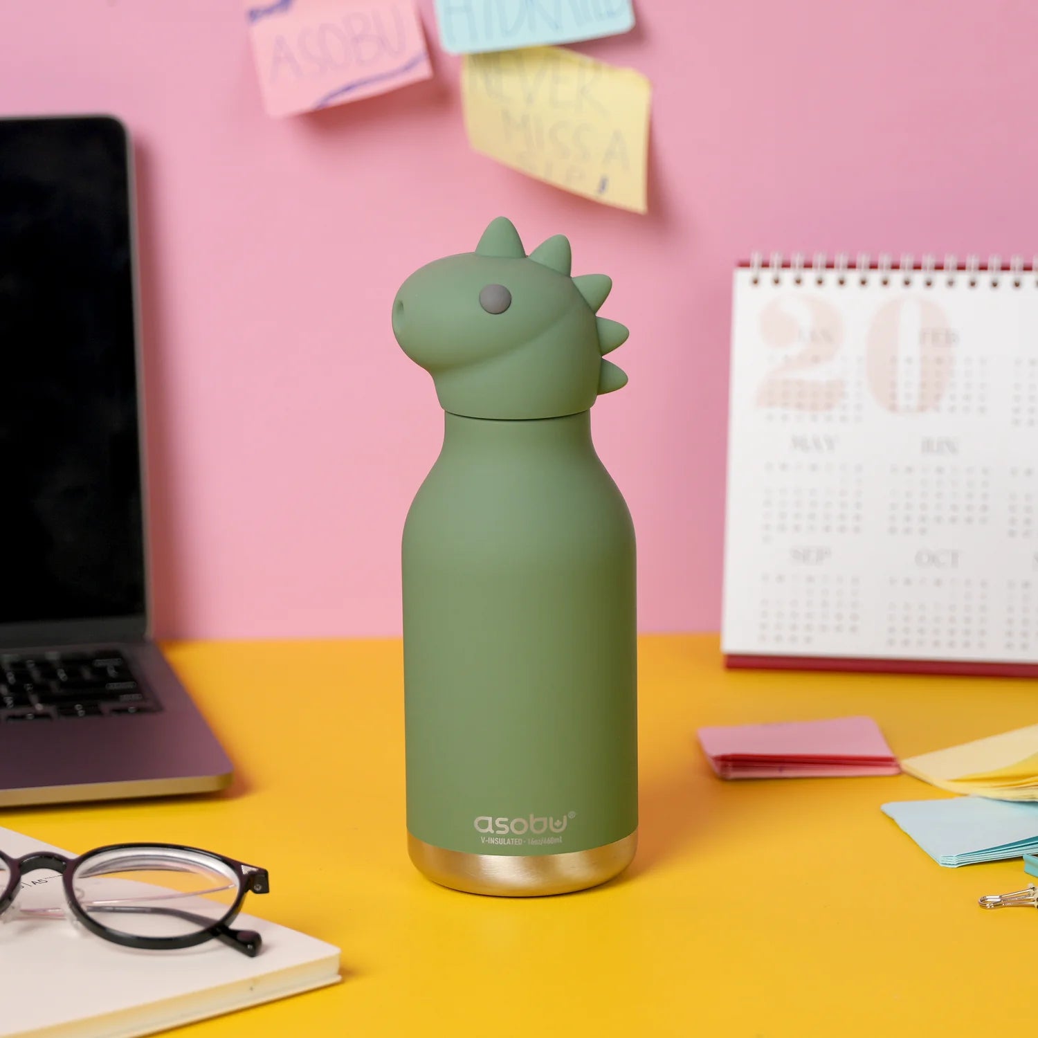 Green dinosaur-shaped water bottle on a yellow desk with a pink wall and laptop in the background.