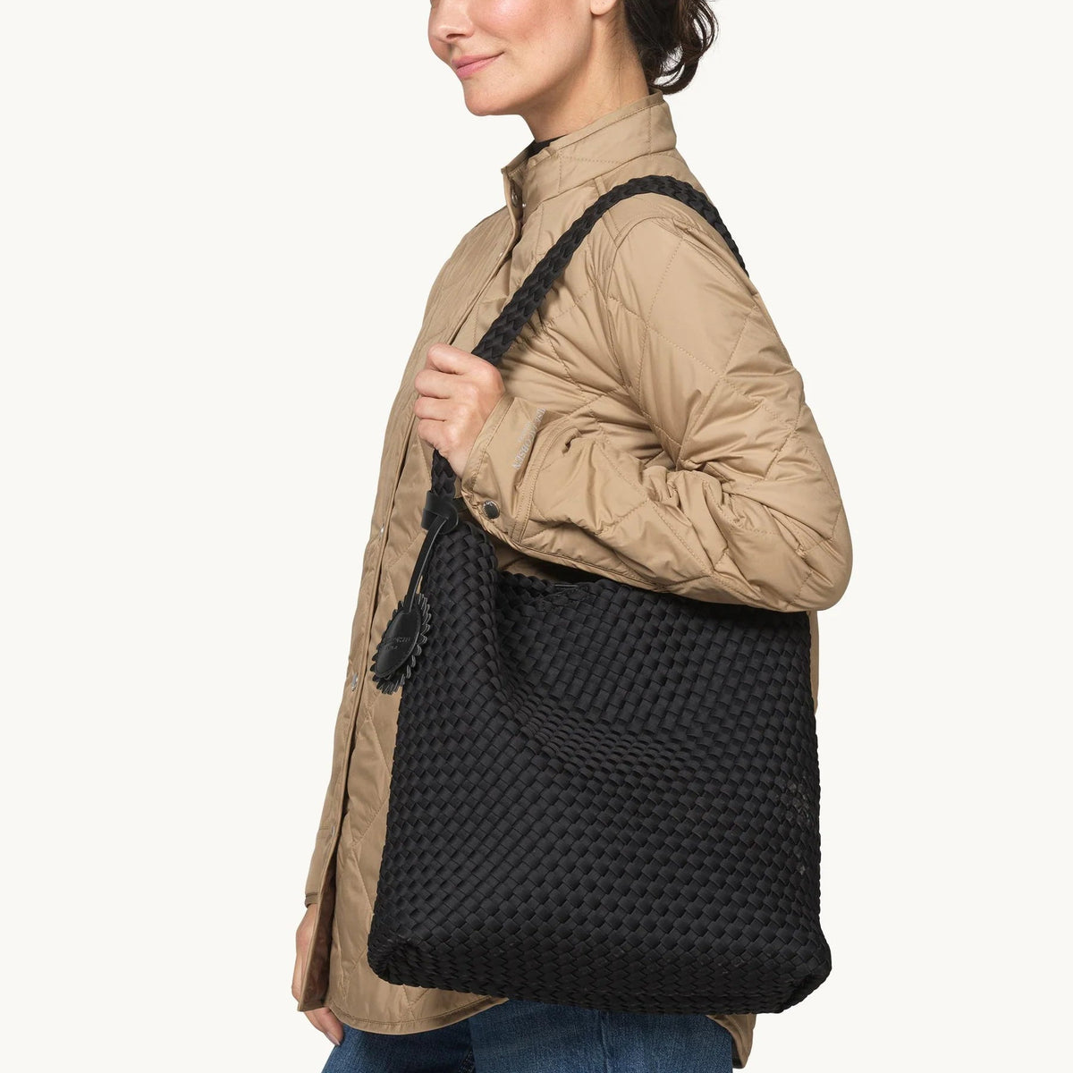 A woman in a tan puffer jacket and blue jeans carries a large black woven tote bag over her shoulder, shown against a plain white background.
