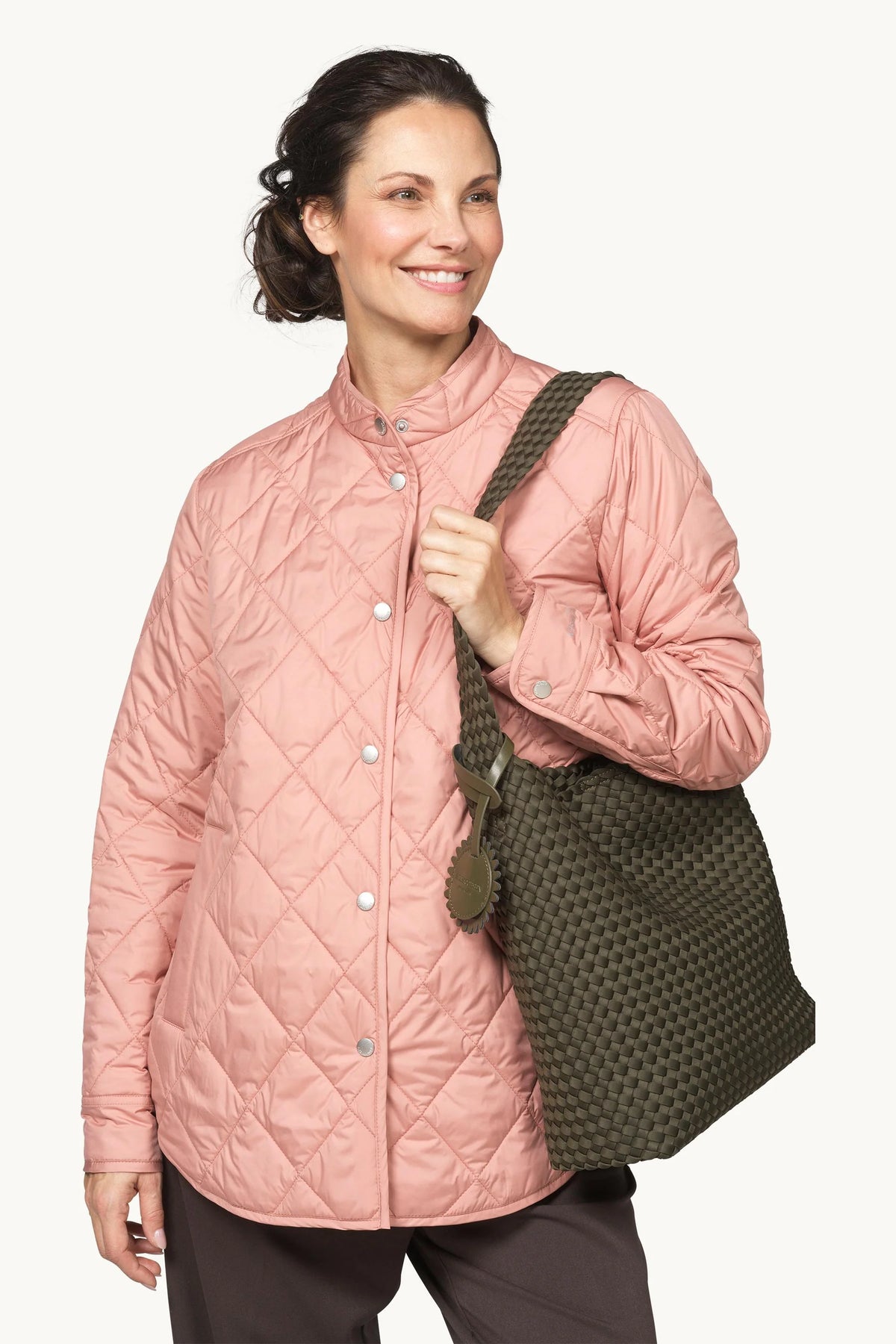A woman wearing a dusty pink diamond-quilted jacket and carrying an olive green woven shoulder bag against a light off-white background.