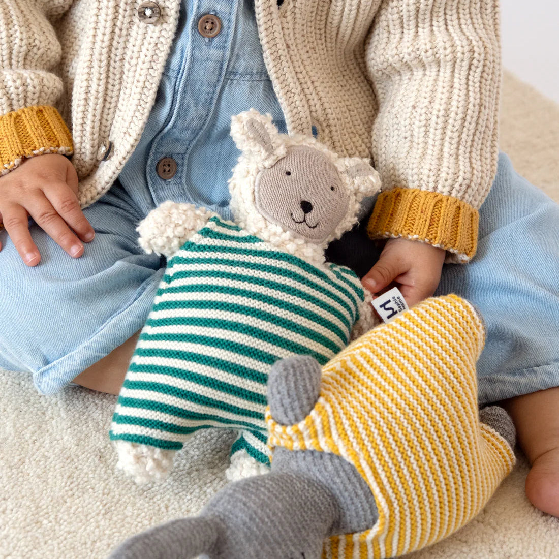 Child wearing a knitted cardigan holding two soft toys on a carpeted floor.