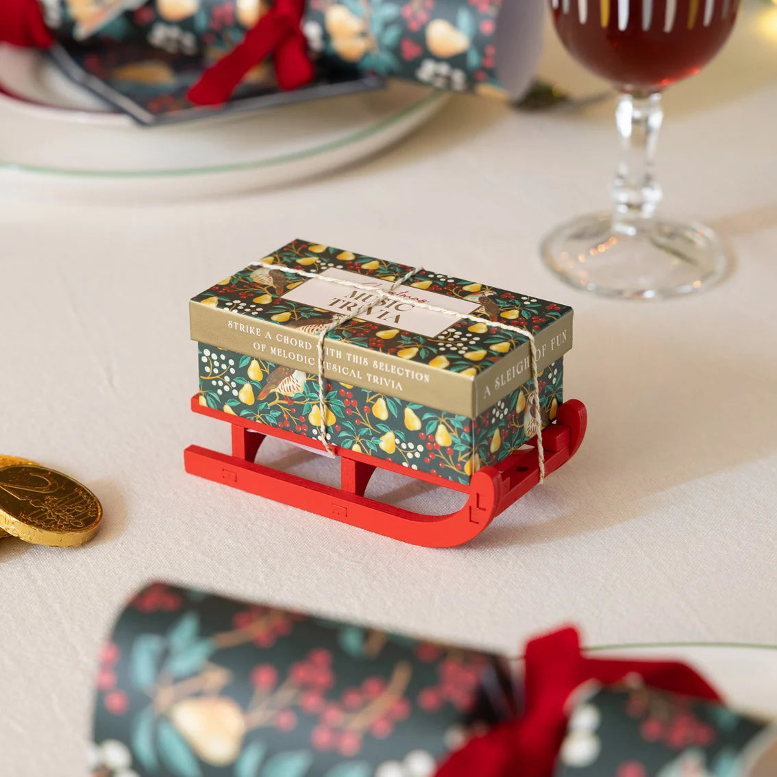 A small gift box wrapped in a festive floral pattern sits on a miniature red sled. The box is tied with a string and has a label that reads 'Merry Gift'.