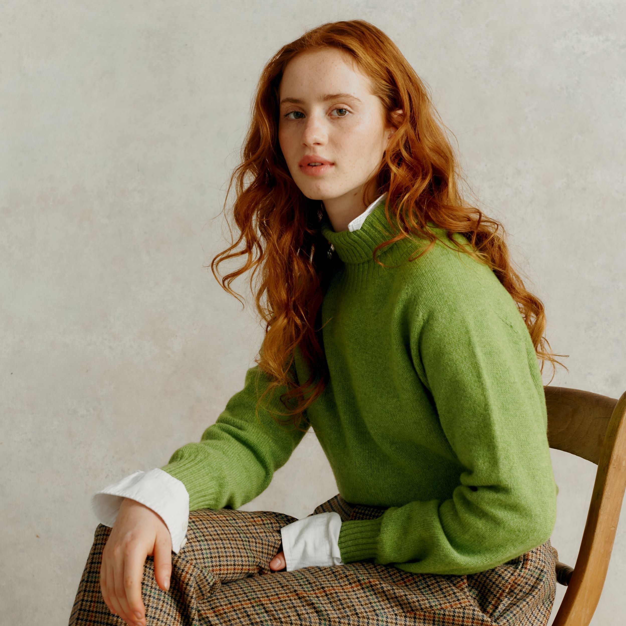 A woman with long red hair sits wearing a light green turtleneck sweater and a brown and beige plaid skirt.