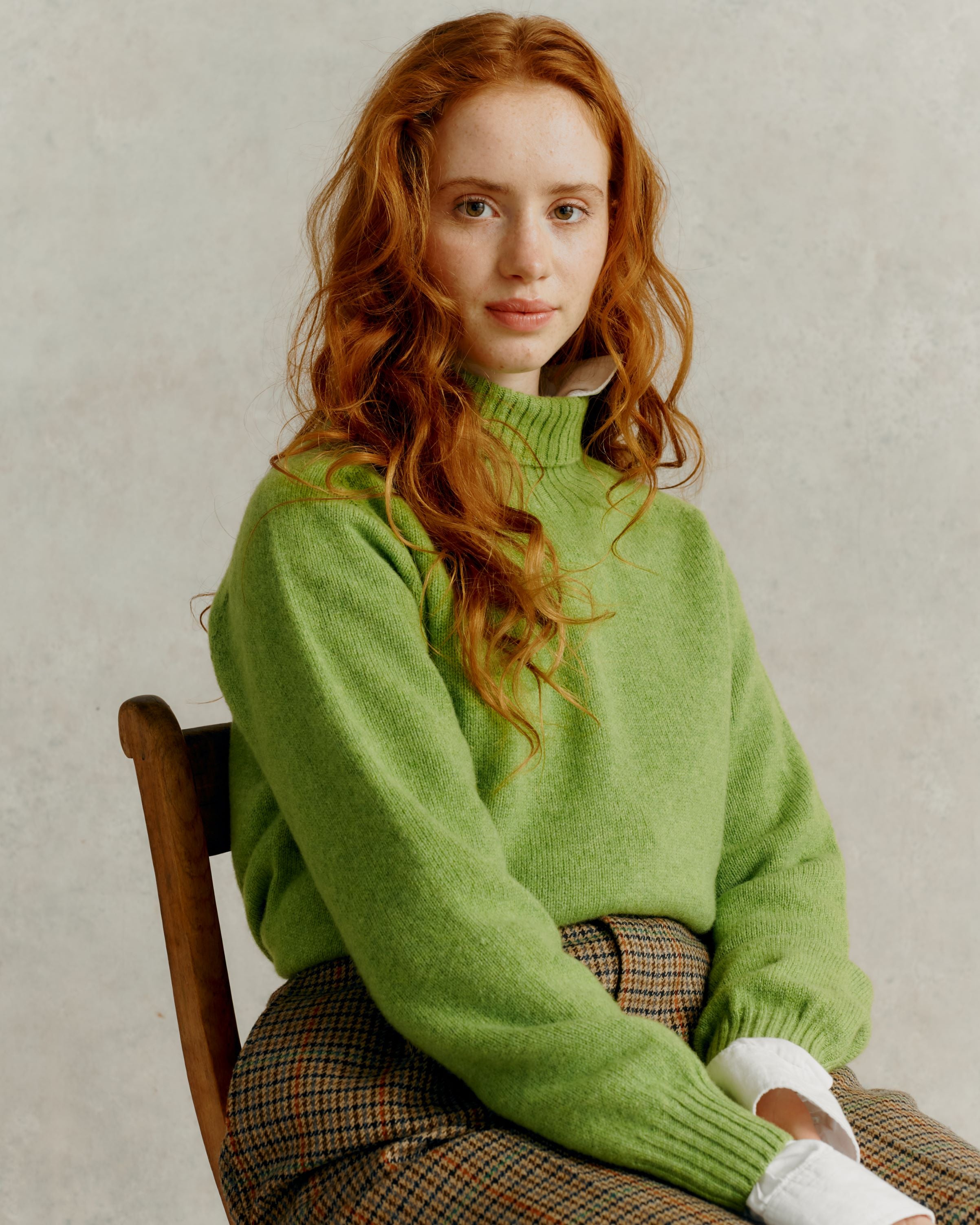 A woman with long red hair sits wearing a light green, high-necked, knit sweater and brown and beige checkered pants.