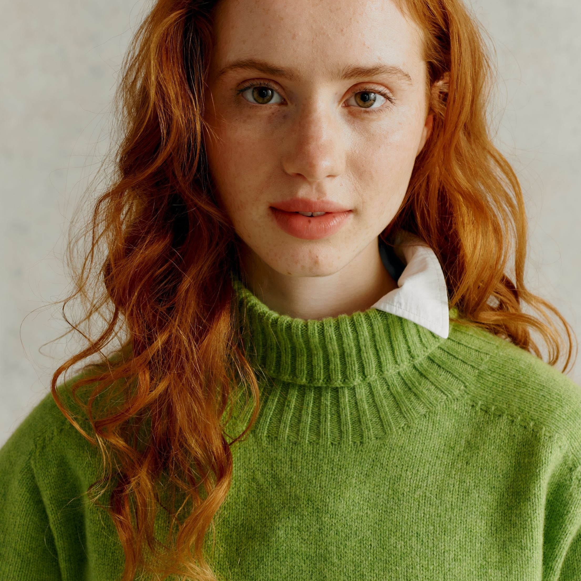 Close-up of a woman with long, curly auburn hair wearing a soft, light green turtleneck sweater.