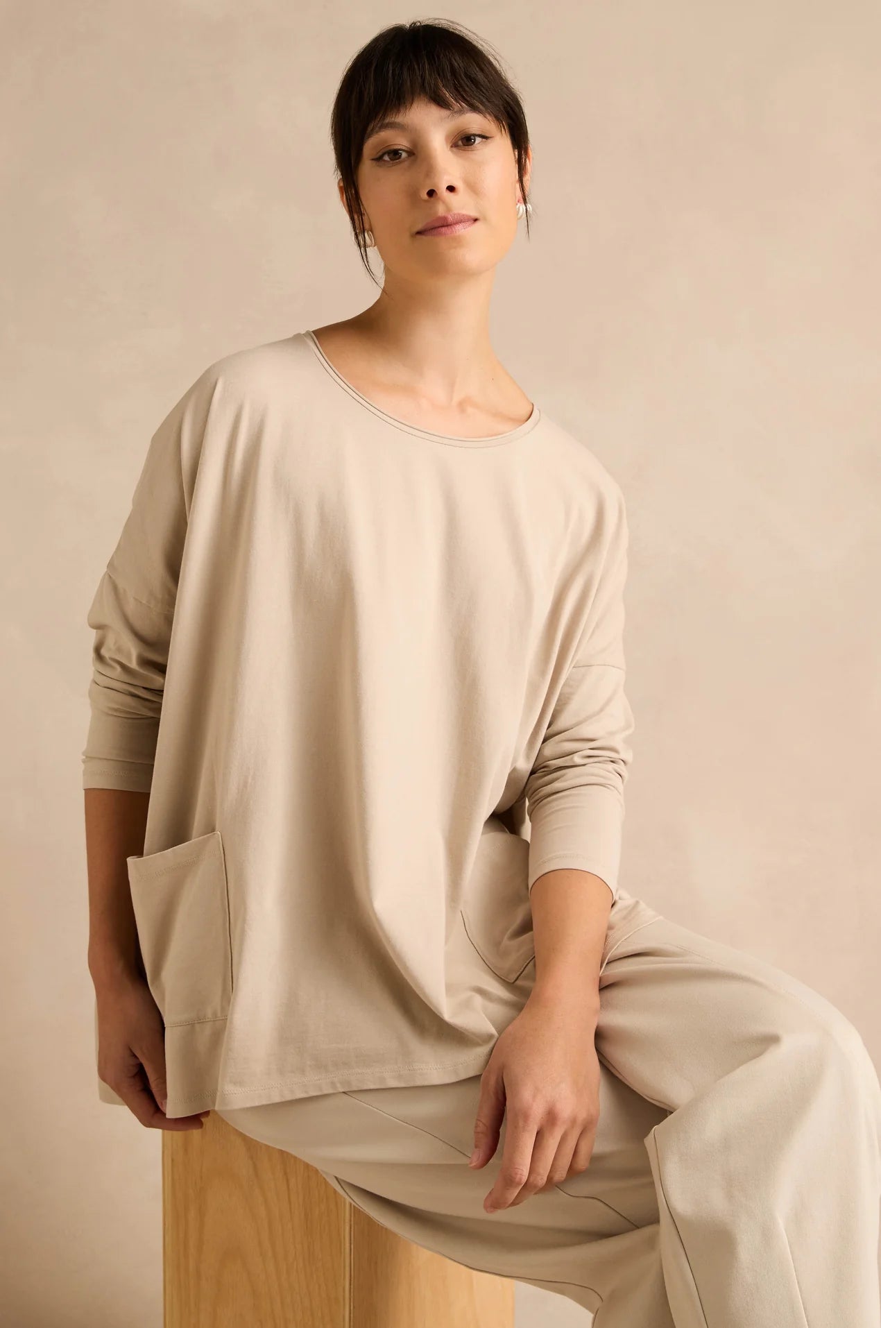 A woman wearing a loose beige long-sleeve top and matching wide-leg pants, seated on a wooden stool against a warm beige studio background.