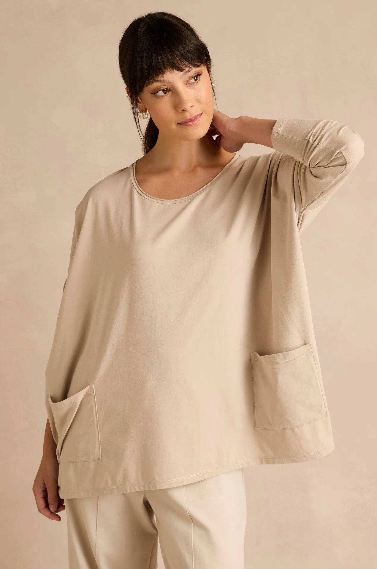 A woman modeling a beige long-sleeve top with large patch pockets, paired with matching beige pants, against a warm beige studio background.