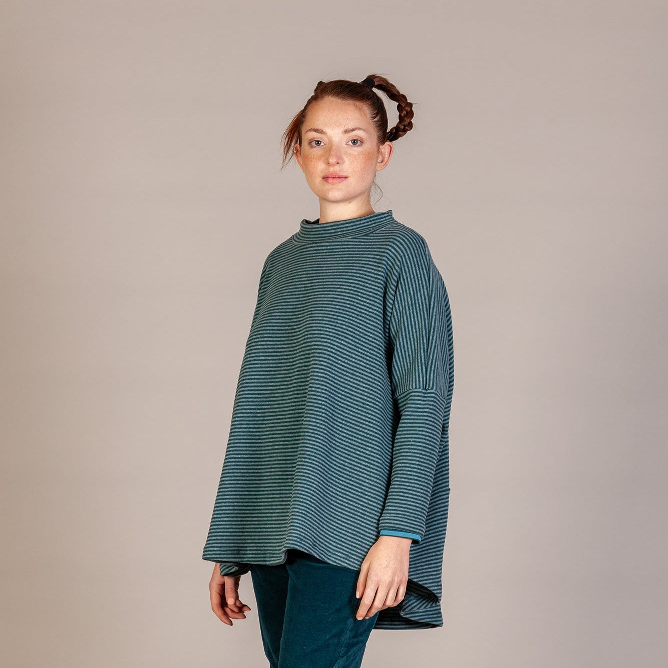 A woman modeling a loose, long-sleeve teal-green ribbed knit top with dark teal pants and brown lace-up boots, standing against a neutral beige studio backdrop.