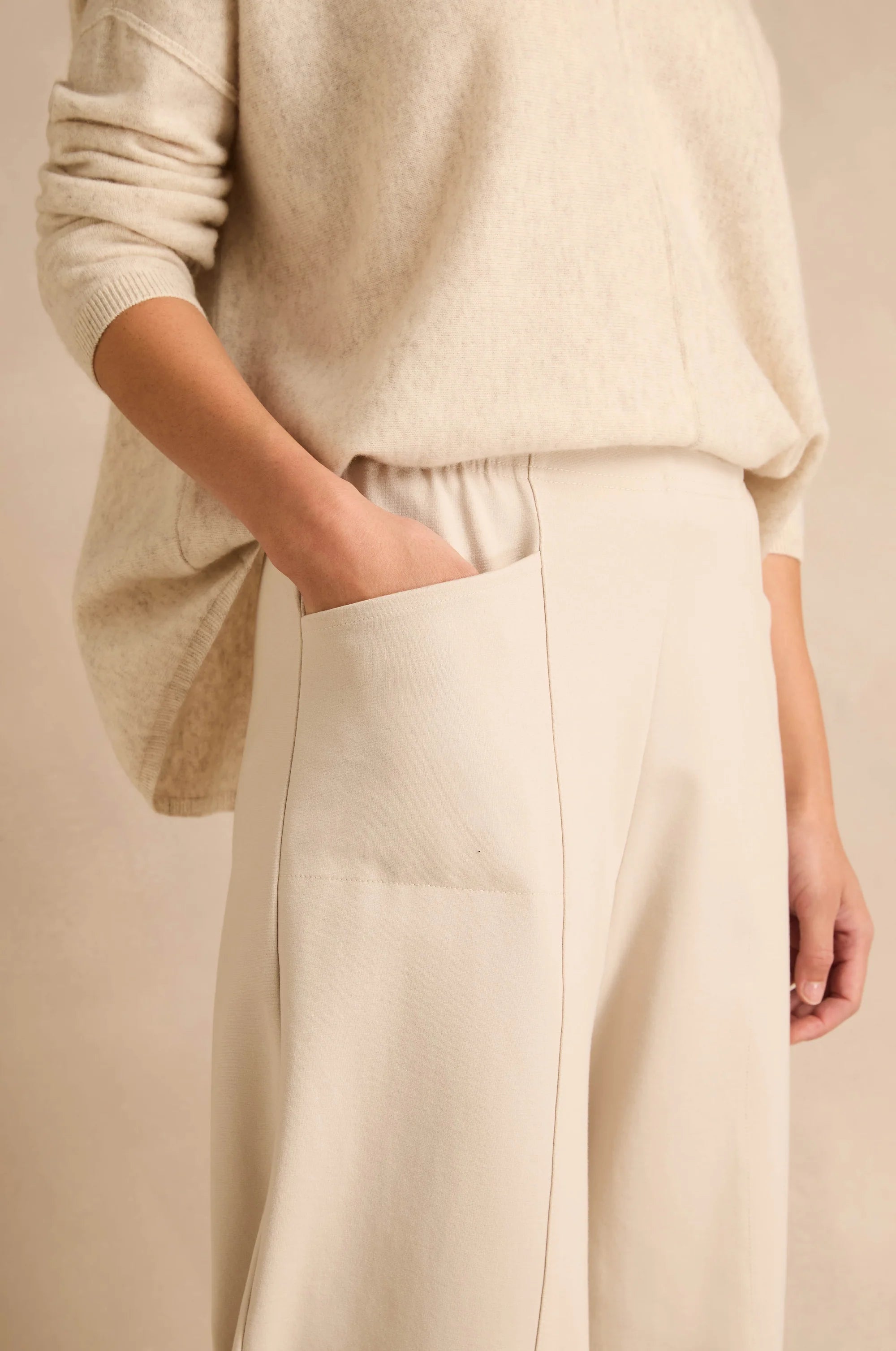 A close-up of a person wearing a cream knit sweater and matching cream wide-leg trousers, with one hand in a trouser pocket, against a soft beige background.