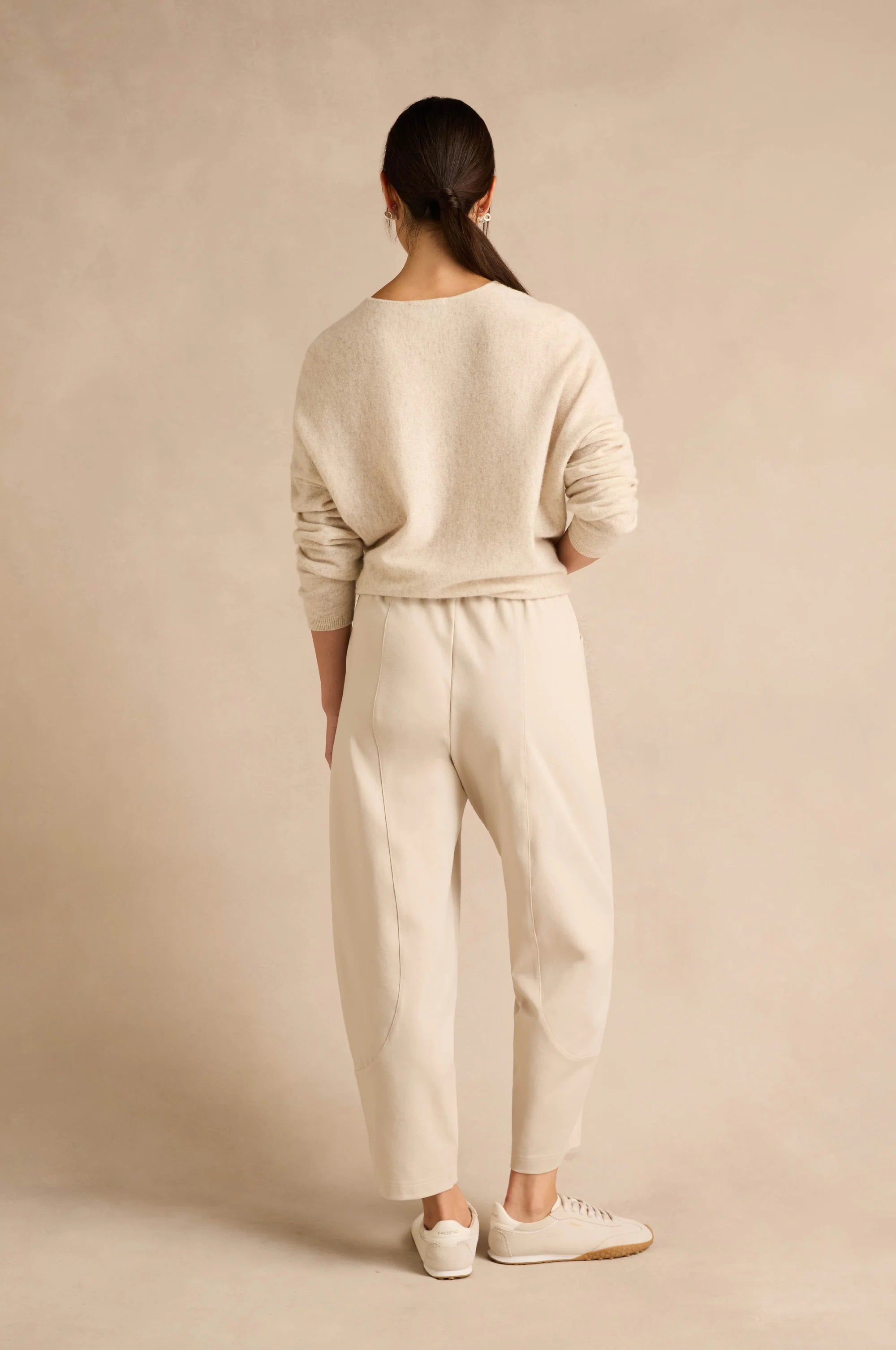 A woman shown from the back wearing a cream-colored knit sweater and matching wide-leg trousers, paired with white sneakers, against a warm beige studio backdrop.