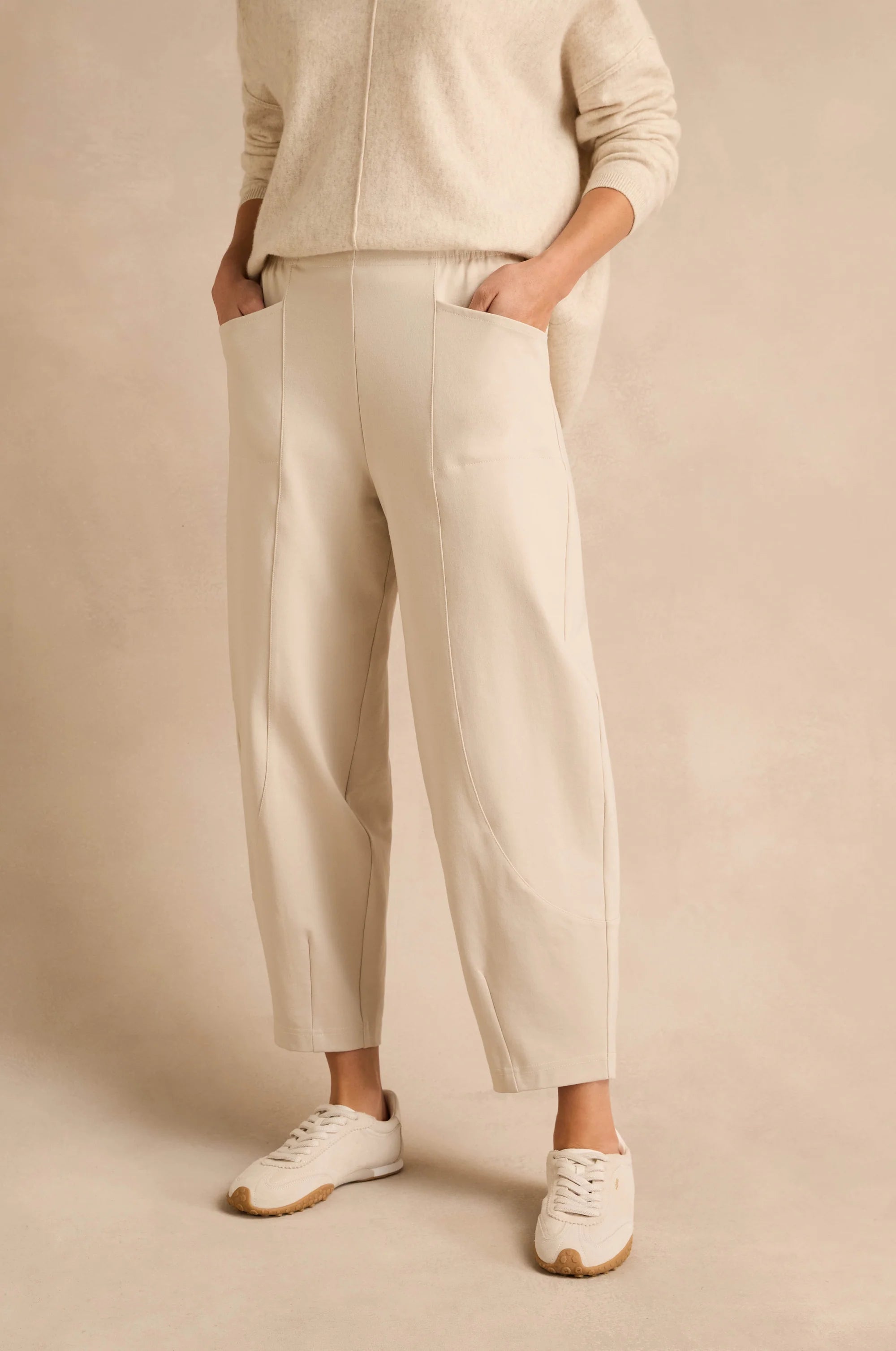 A person wearing cream-colored wide-leg cropped trousers with side pockets, paired with a matching cream knit top and white sneakers, standing against a warm beige studio backdrop.
