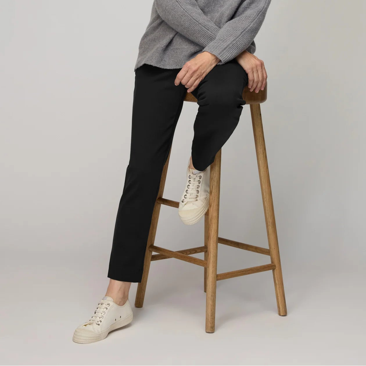 Woman sitting on a wooden stool wearing a gray sweater and black pants against a plain background