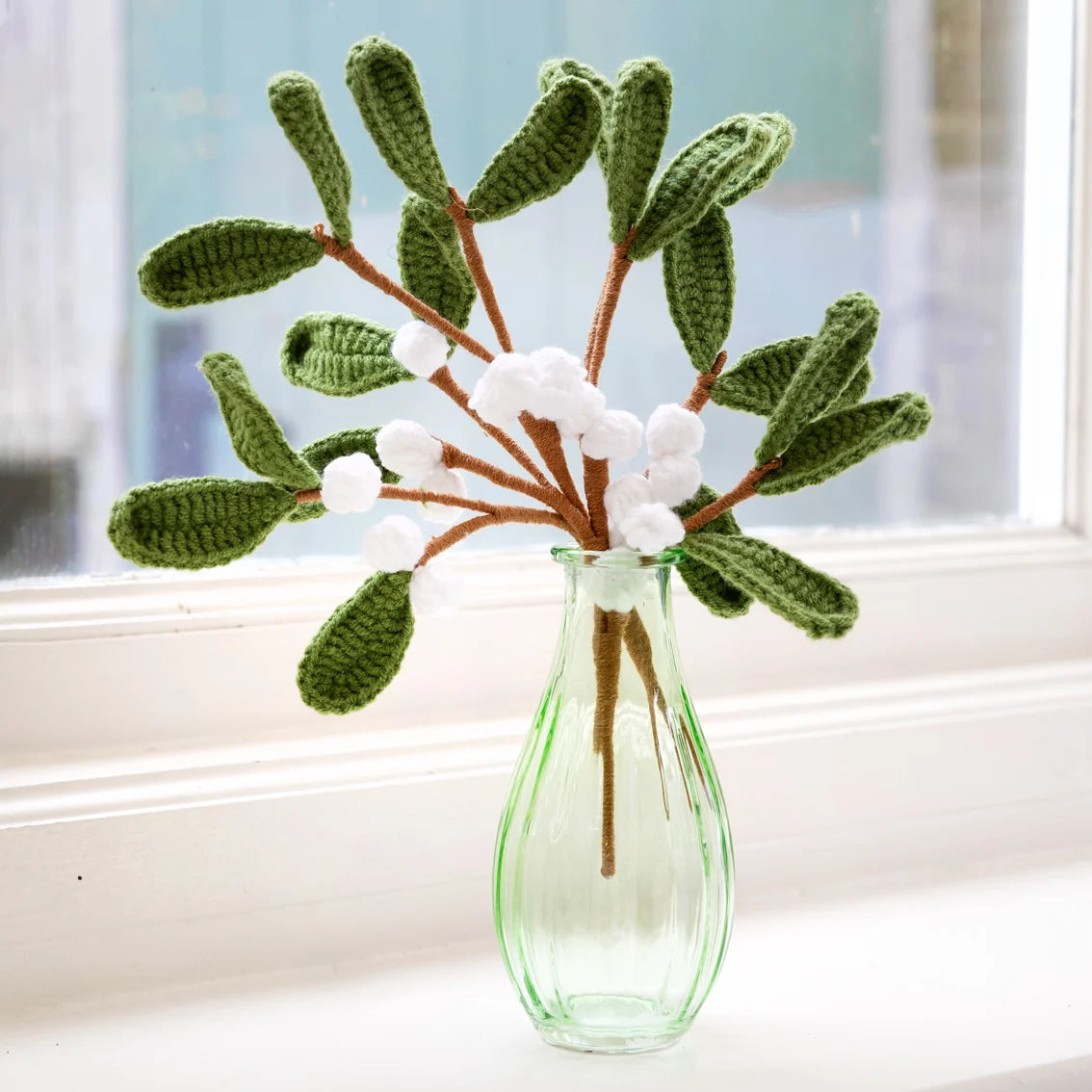 Crocheted green leaves with white berries in a clear glass vase on a windowsill.