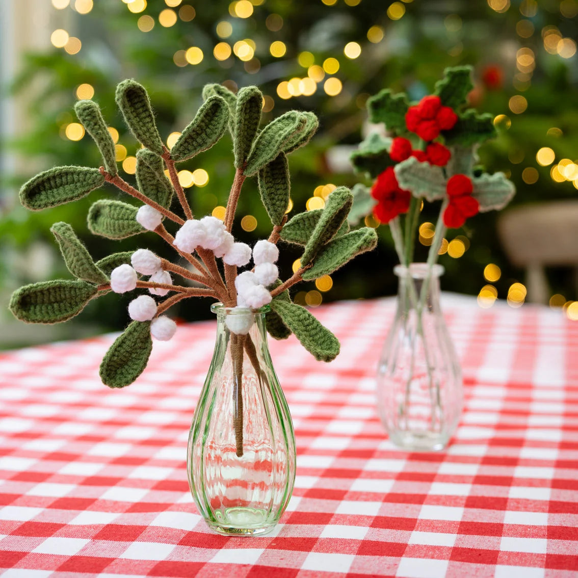 Handmade crocheted mistletoe branches with green leaves and white berries in a small clear glass vase, set on a red and white checkered tablecloth with a blurred Christmas tree and lights in the background.